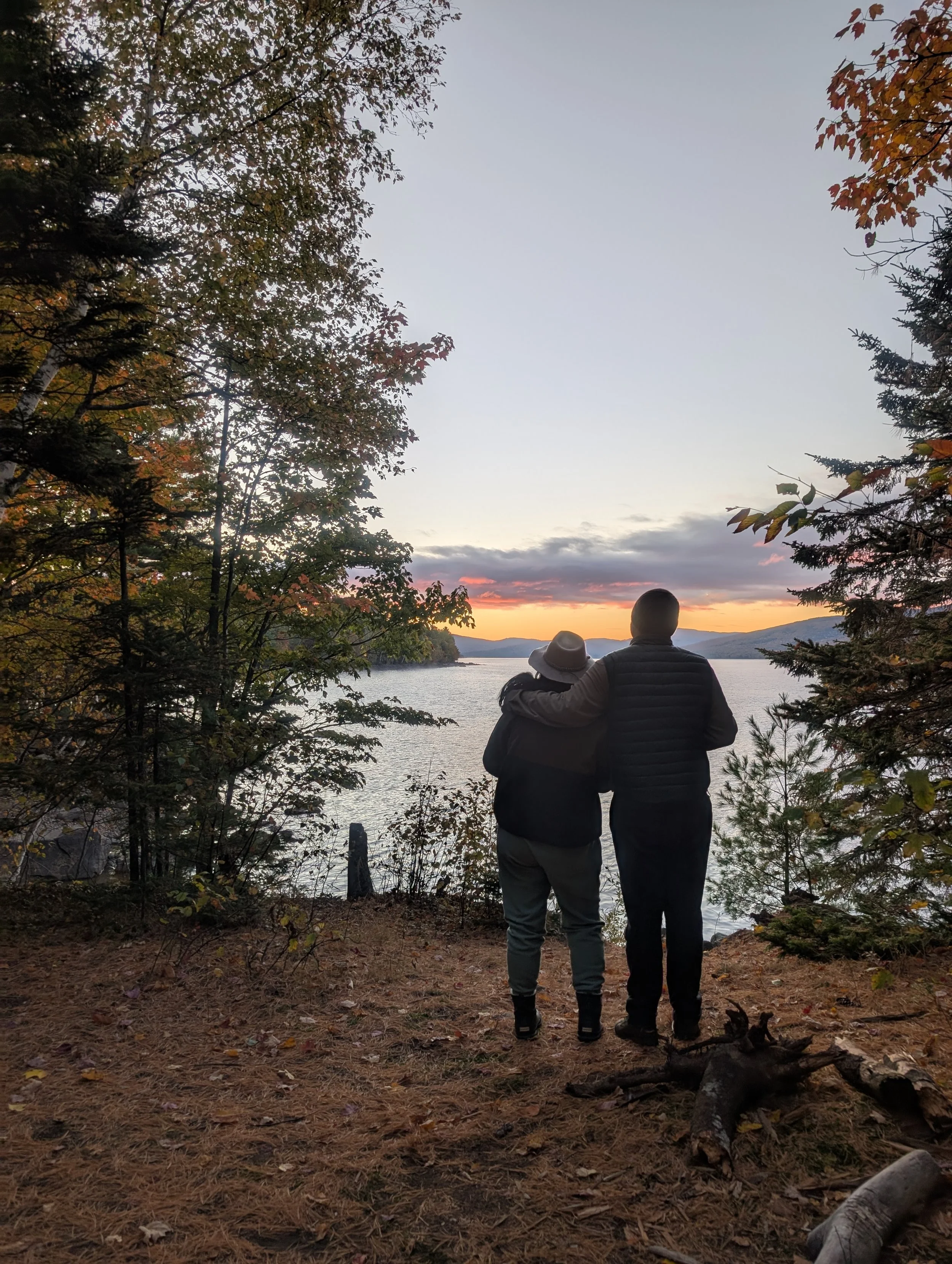 Two people standing close together by a lake at sunset, framed by trees with autumn leaves, embracing and looking out at the water and mountains in the distance.