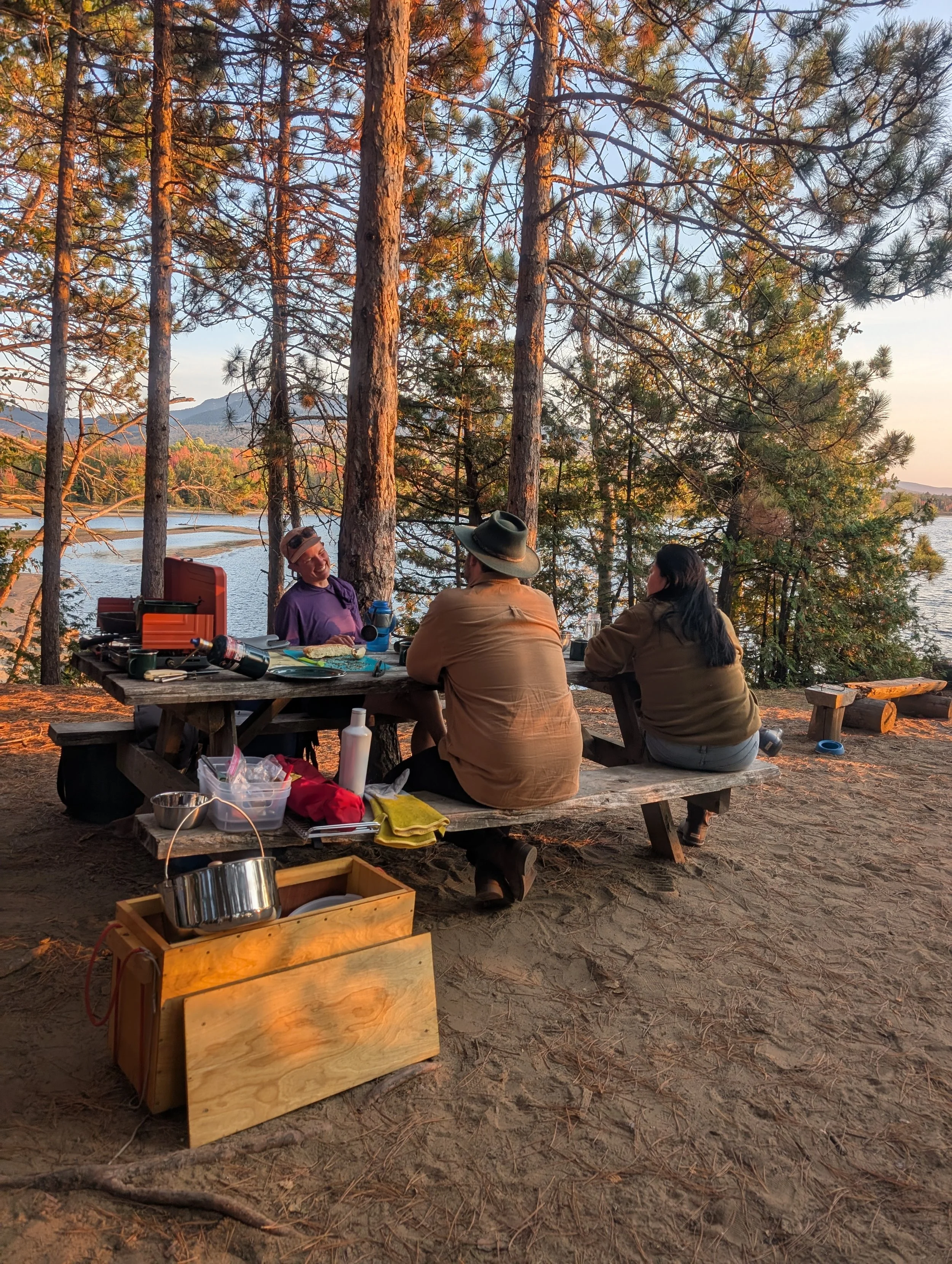 Four people are sitting around a wooden picnic table outdoors in a forested area near a lake. There are trees with autumn leaves, and mountains are visible in the background. The table has various items like water bottles, cups, and a backpack. The s