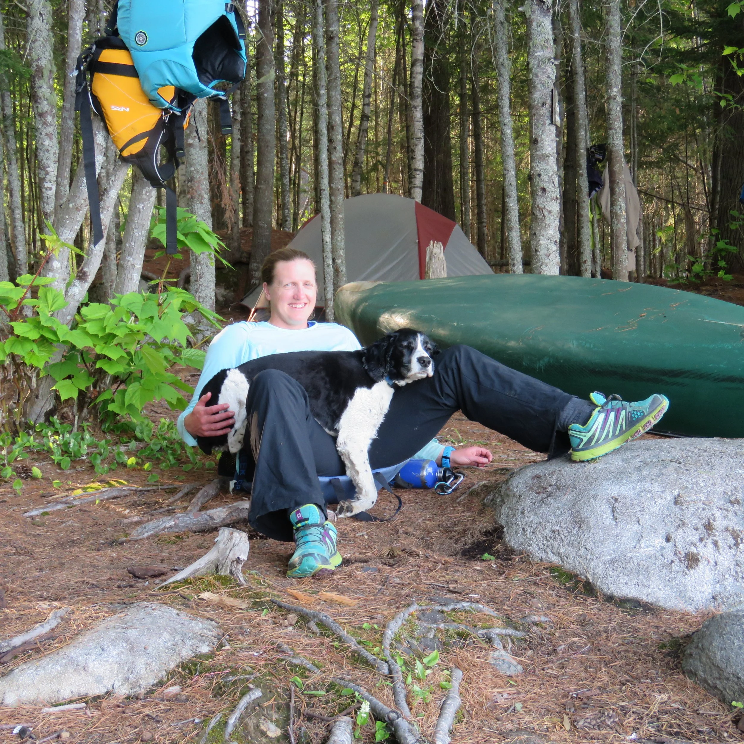 A person sitting outdoors in a forested area, leaning back on their hands, with two black and white dogs resting on their lap, near a green canoe, surrounded by trees, a tent, and camping gear.