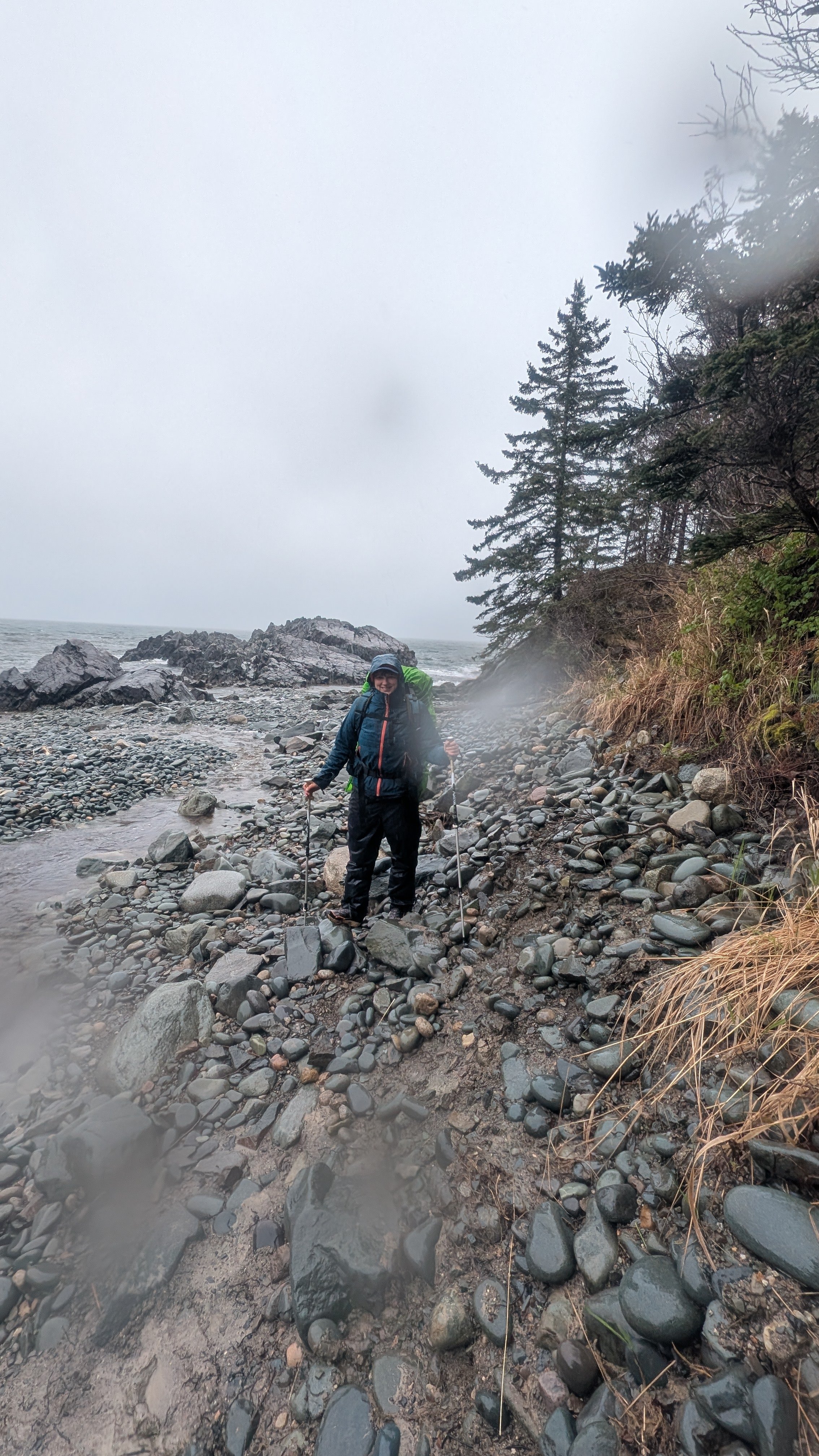 Bold Coast Trail Cutler, Maine. A person in outdoor hiking gear with trekking poles standing on a rocky beach near the water and trees under an overcast sky.