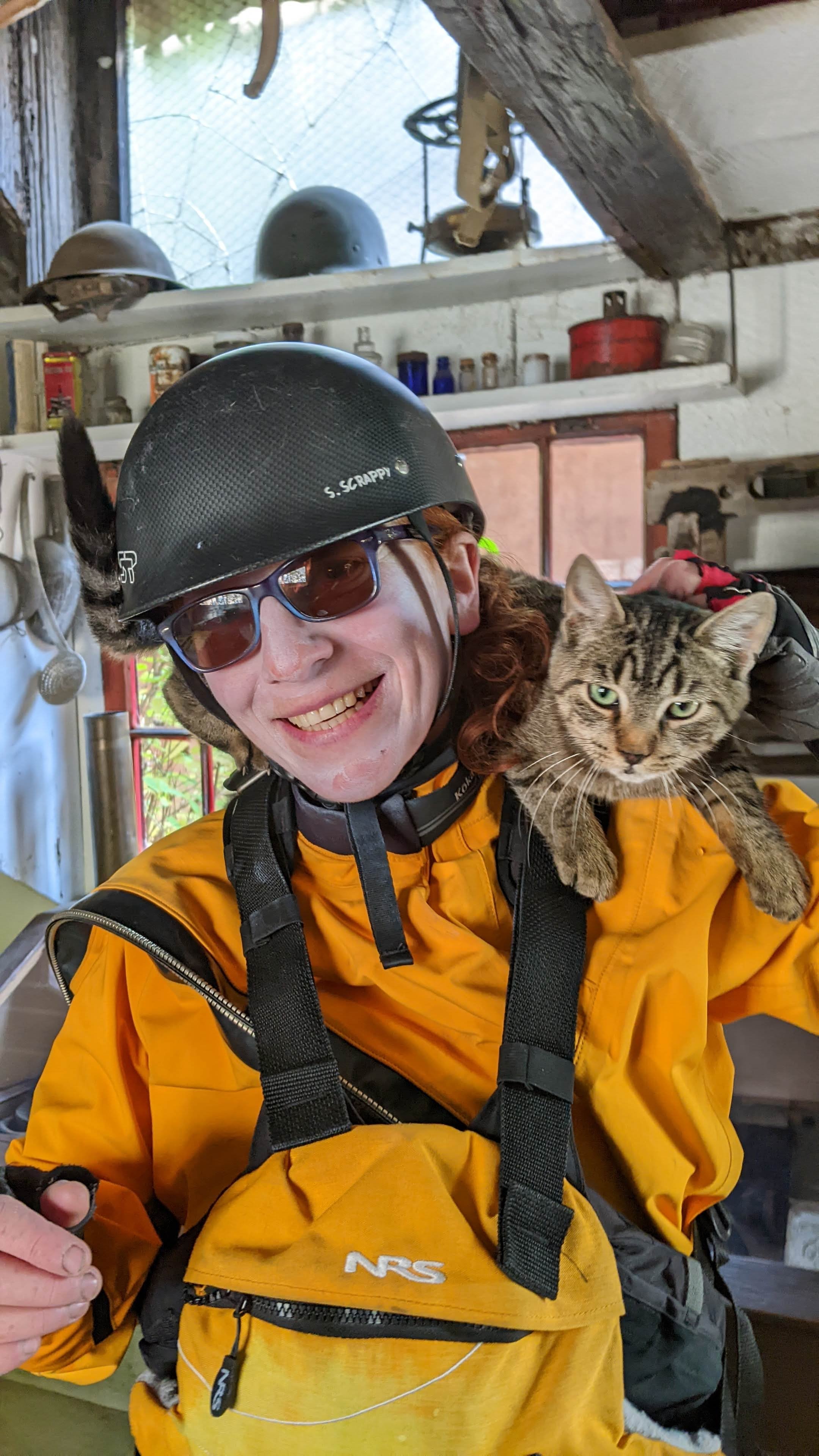 Fall canoeing on the Salmon River, ID. A person wearing a yellow dry suit, black helmet, and glasses is smiling while holding a tabby cat with green eyes on their shoulder. The background shows a rustic indoor space with shelves, helmets, and cans.