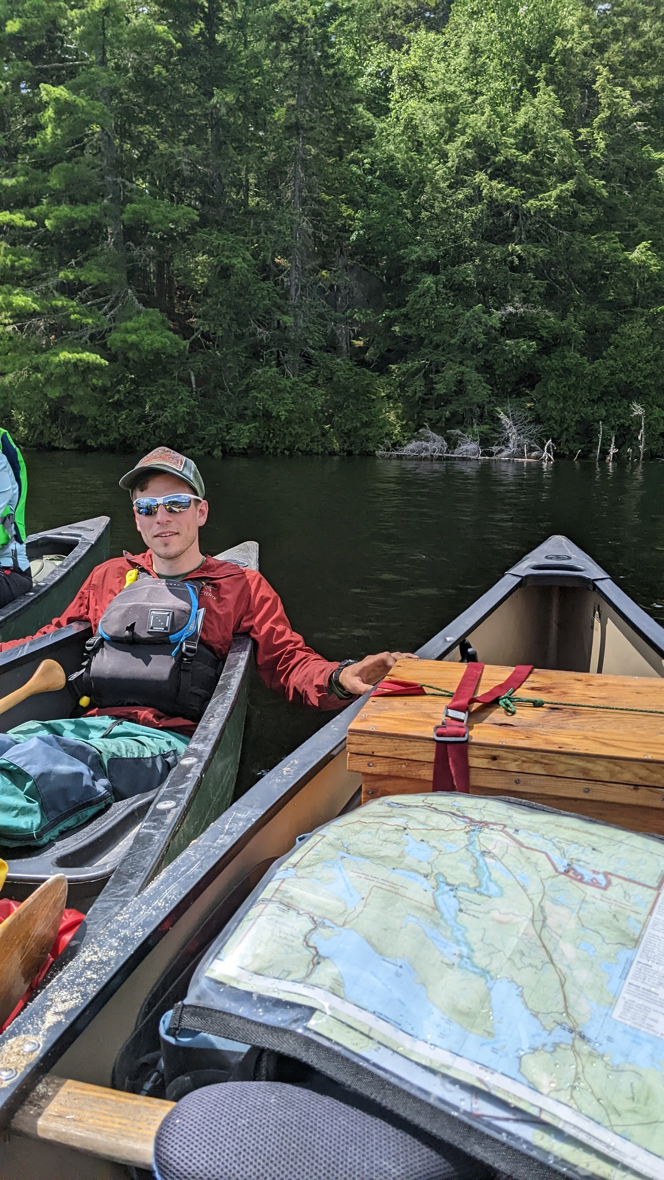 Passy Lake, Maine. A man sitting in a canoe on a river among green trees, with a box and a map in the foreground.