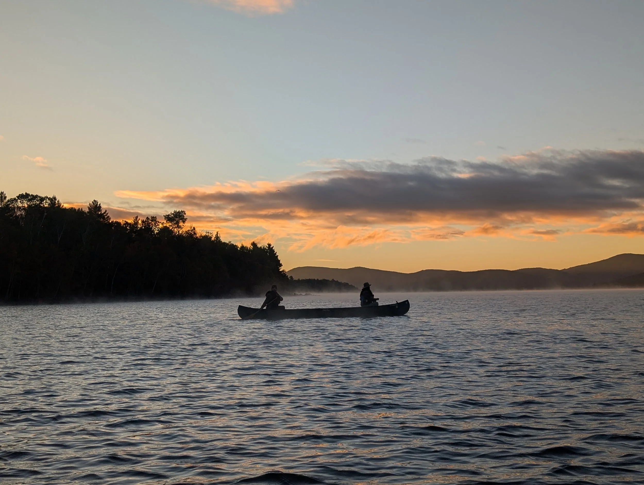 Flagstaff Lake, Maine. A serene scene of two people in a canoe on a calm lake during sunset, with a tree-lined shoreline and mountains in the background, and a sky with clouds and a hint of orange and pink hues.