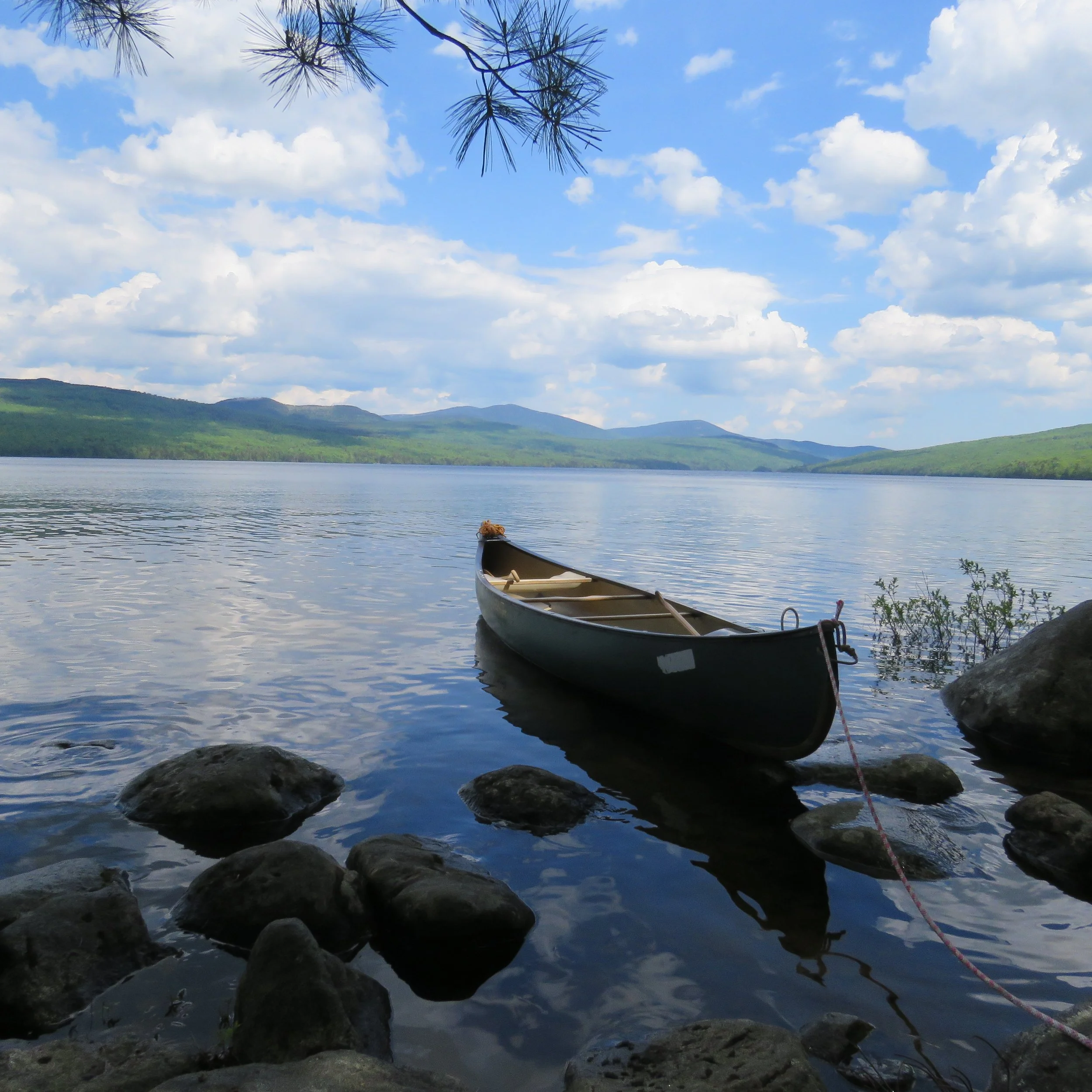 Richardson Lake, Maine. A peaceful lake surrounded by green hills and mountains under a blue sky with scattered clouds, with a canoe resting on the rocky shoreline.