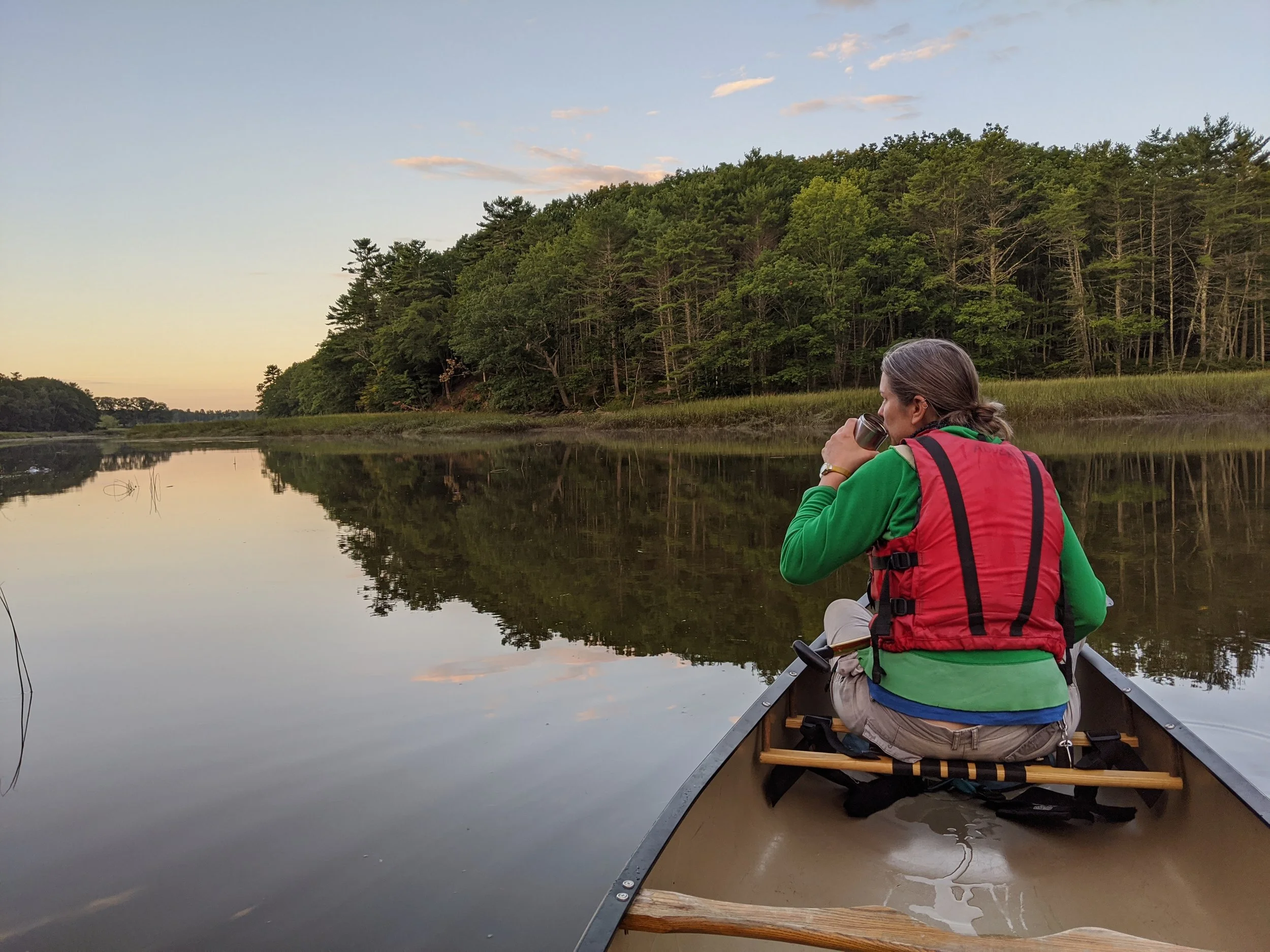 A person in a red vest and green hoodie is sitting in a canoe on a calm river, drinking from a stainless steel cup, with a dense forest of green trees and a partly cloudy sky reflected in the water.