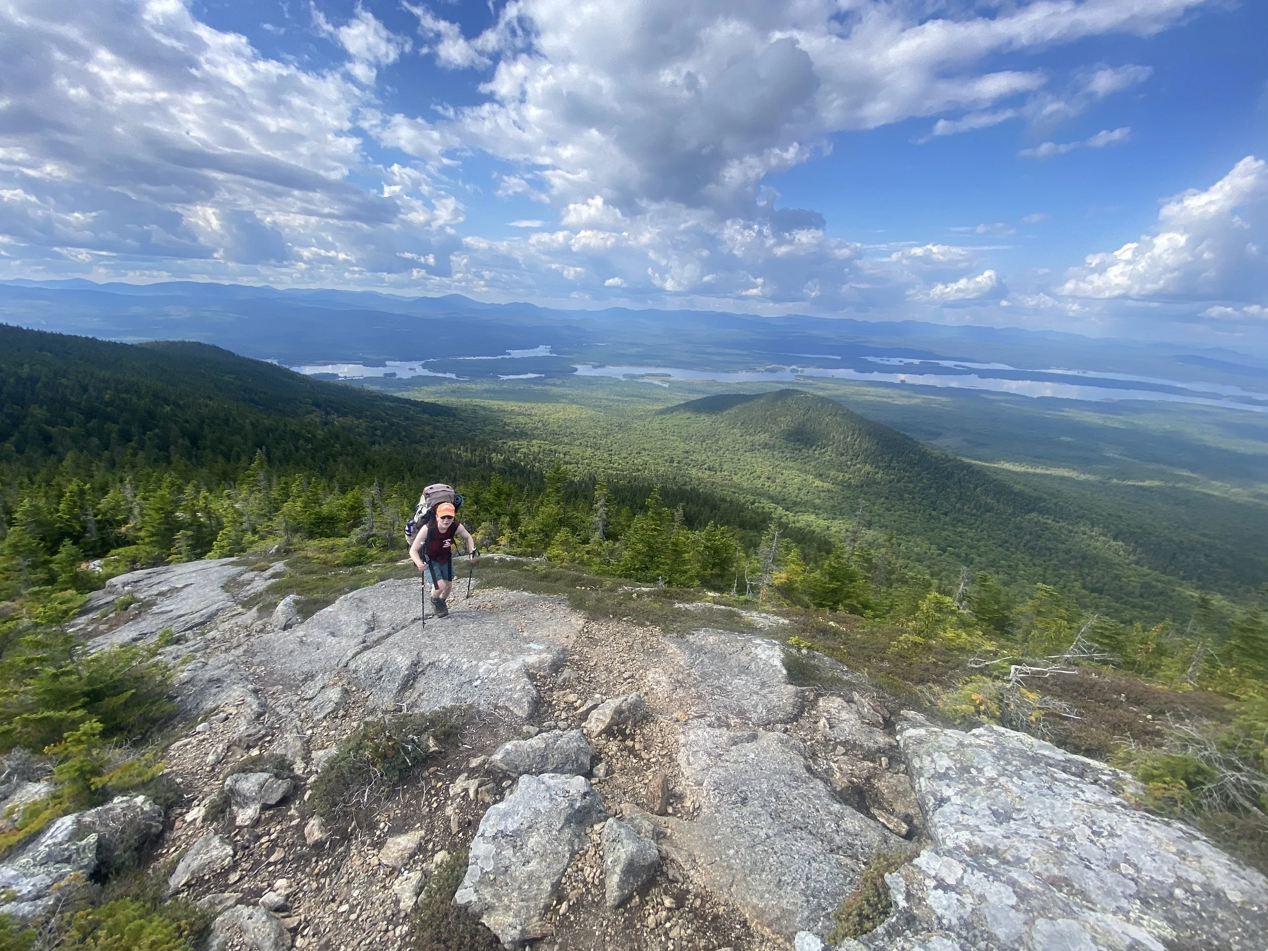 Bigelow Range, Maine. A person hiking up a rocky mountain trail with a scenic view of a vast green landscape, river lakes, and distant mountains under a partly cloudy sky.
