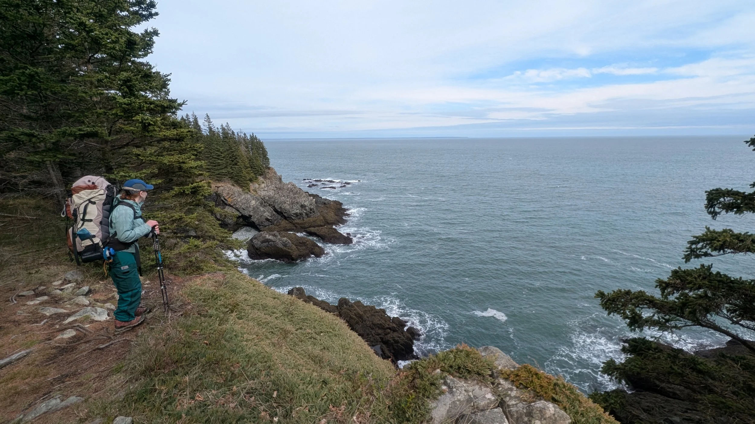Bold Coast Trail Cutler, Maine. Hiker with a large backpack and trekking poles standing on a rocky grassy coastline, looking out over the ocean with trees and rocks along the shore and a partly cloudy sky.