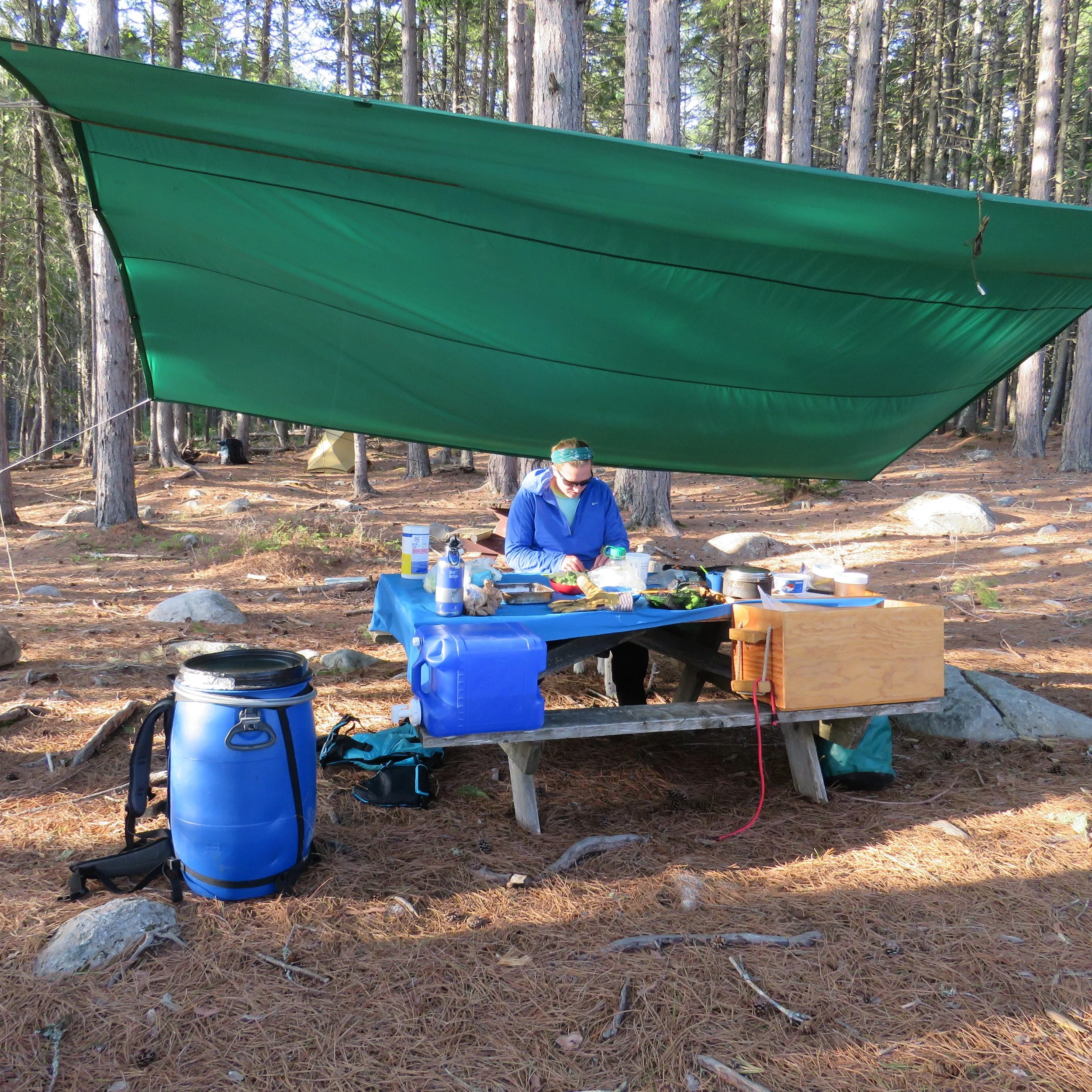 Richardson Lake, Maine. A person wearing a blue jacket and sunglasses sitting at an outdoor table under a green tarp in a forest.