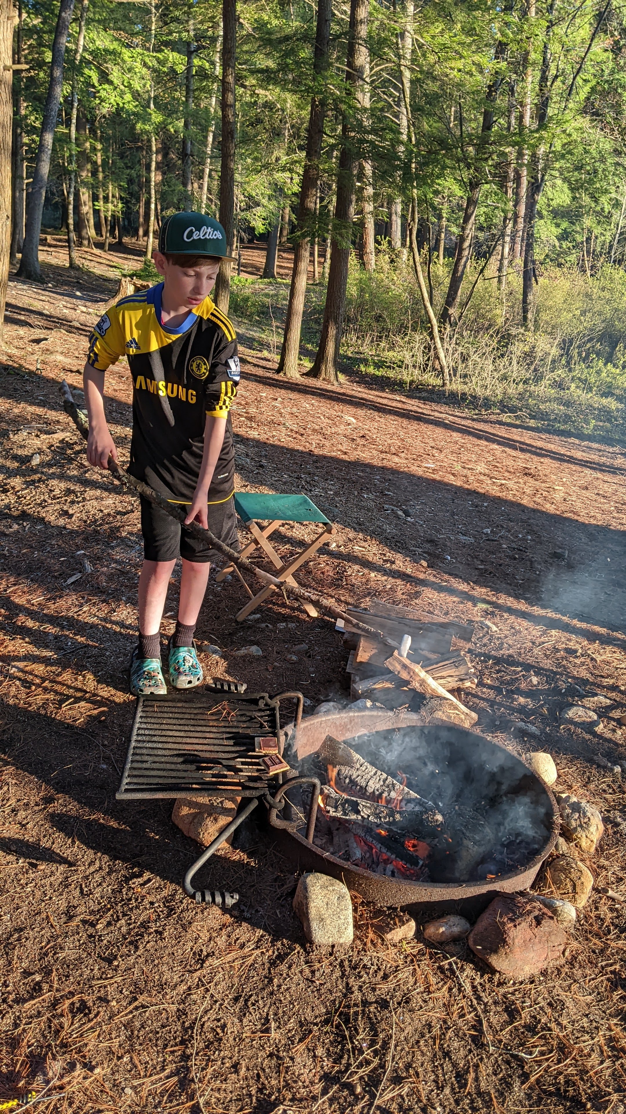 A young boy in a Chelsea football jersey and a baseball cap standing near a campfire, cooking food on a grill in a forested area with trees and sunlight filtering through.