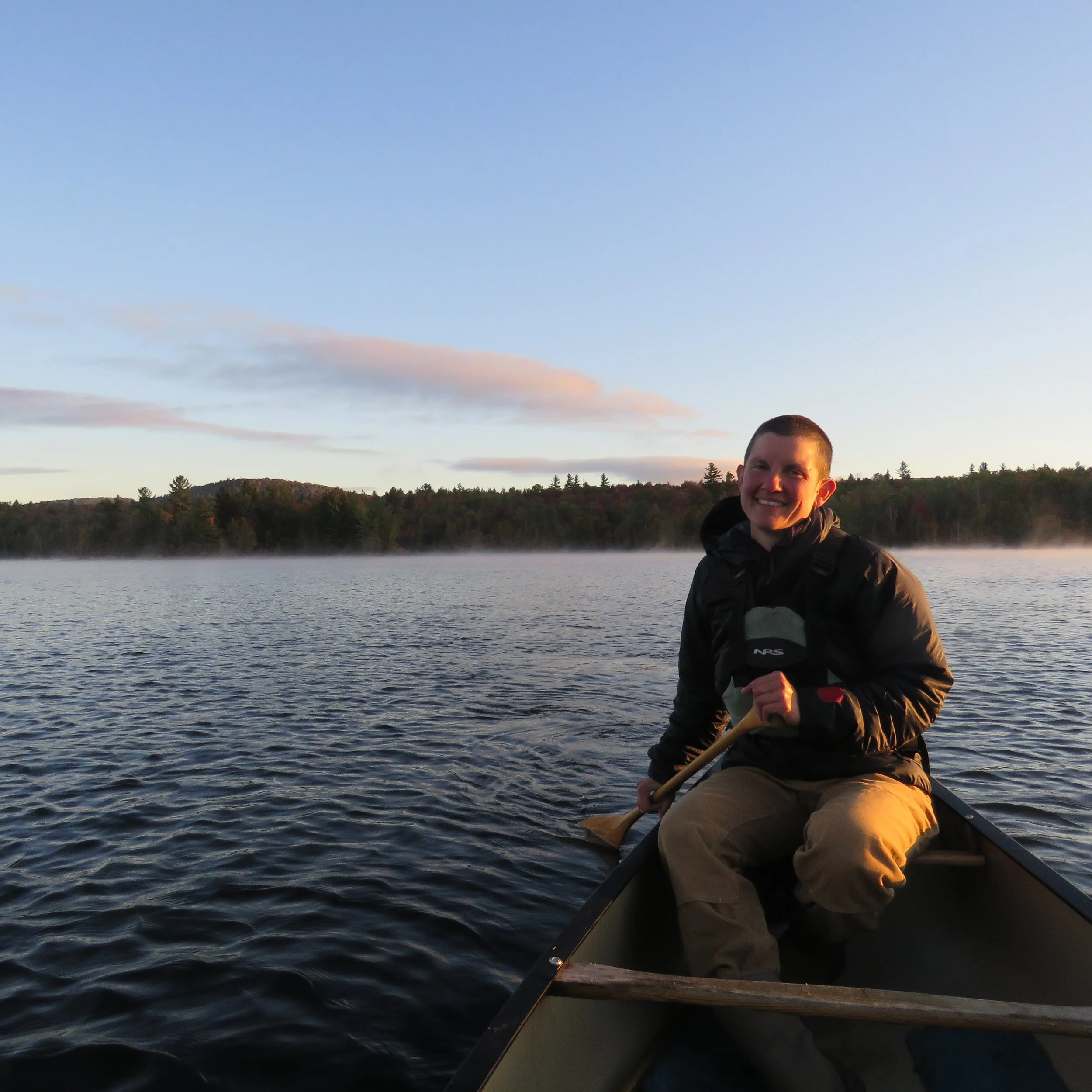 Fall canoeing on Flagstaff Lake, Maine. A person smiling while paddling a canoe, on a Lake during sunrise, with trees and hills in the background. The mist rises off the water.