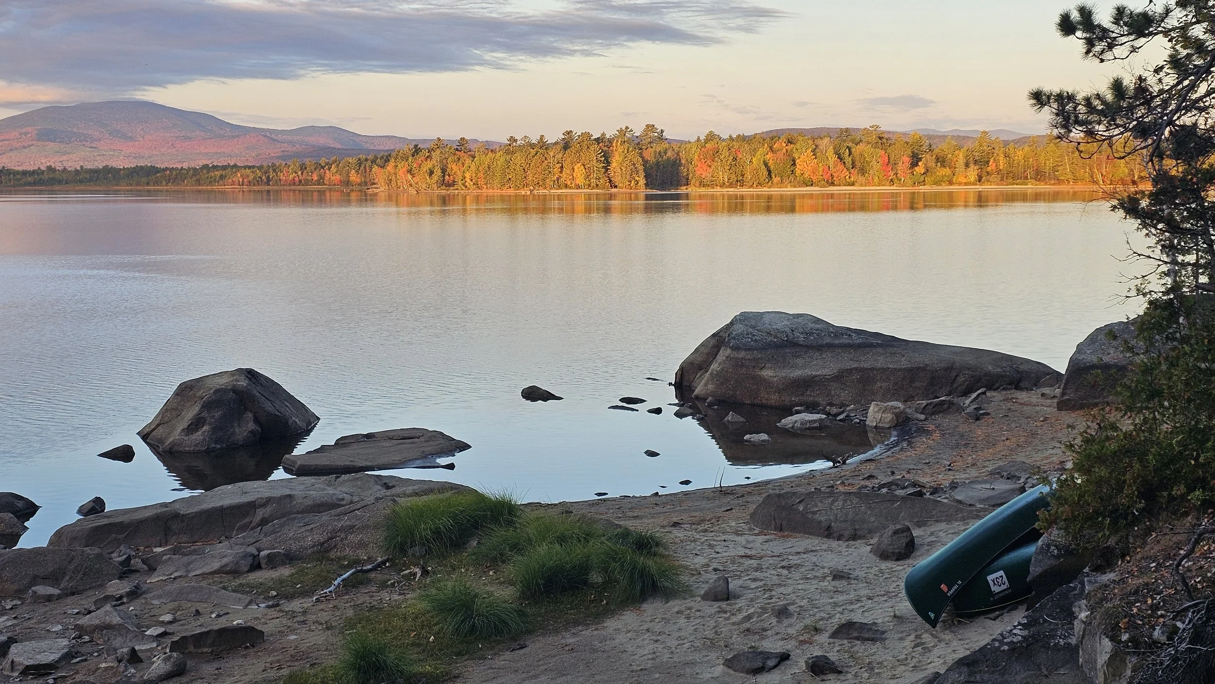 Flagstaff Lake, Maine. A peaceful lake scene at sunset with calm water, a rocky shore in the foreground, trees with fall foliage across the lake, and mountains in the distance under a partly cloudy sky.