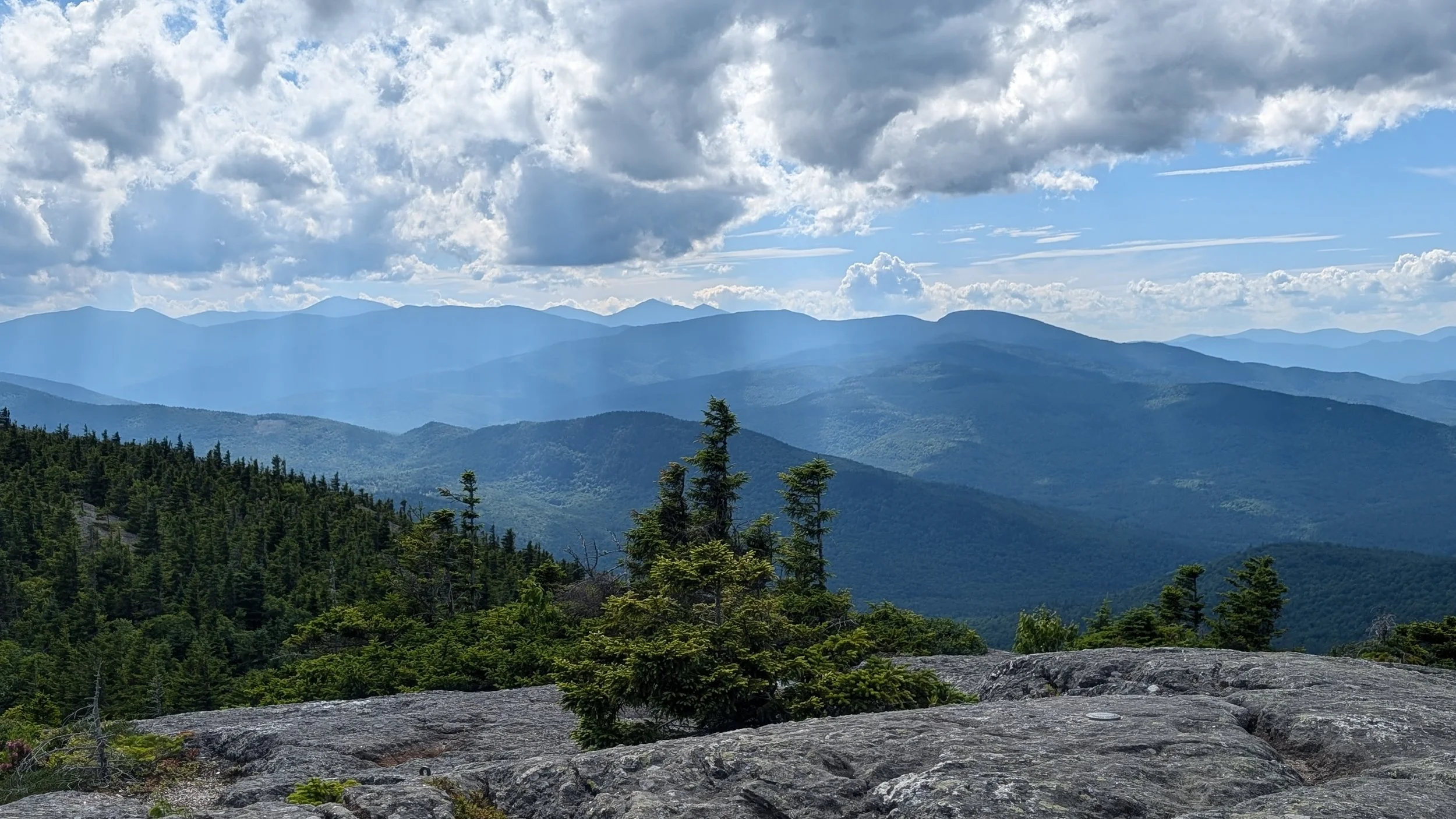Caribou Speckled Wilderness, Maine. Mountain range with green forest in the foreground, layers of blue mountains in the background, and a partly cloudy sky.