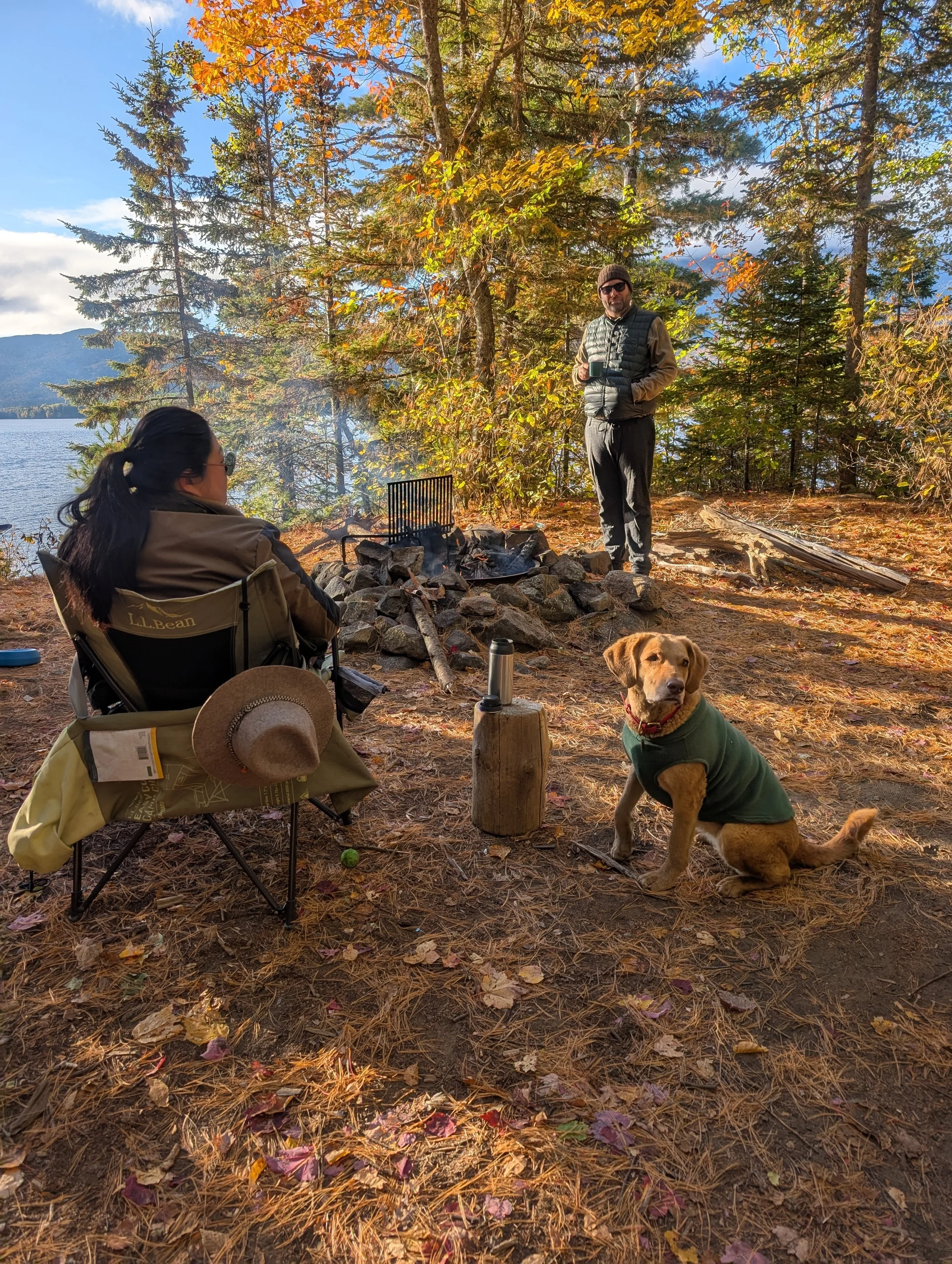 A man and a woman enjoying a campfire by a lake surrounded by trees with autumn foliage. The man is standing holding a mug, while the woman is sitting in a camping chair. A golden retriever dog wearing a green sweater is sitting nearby. The ground is