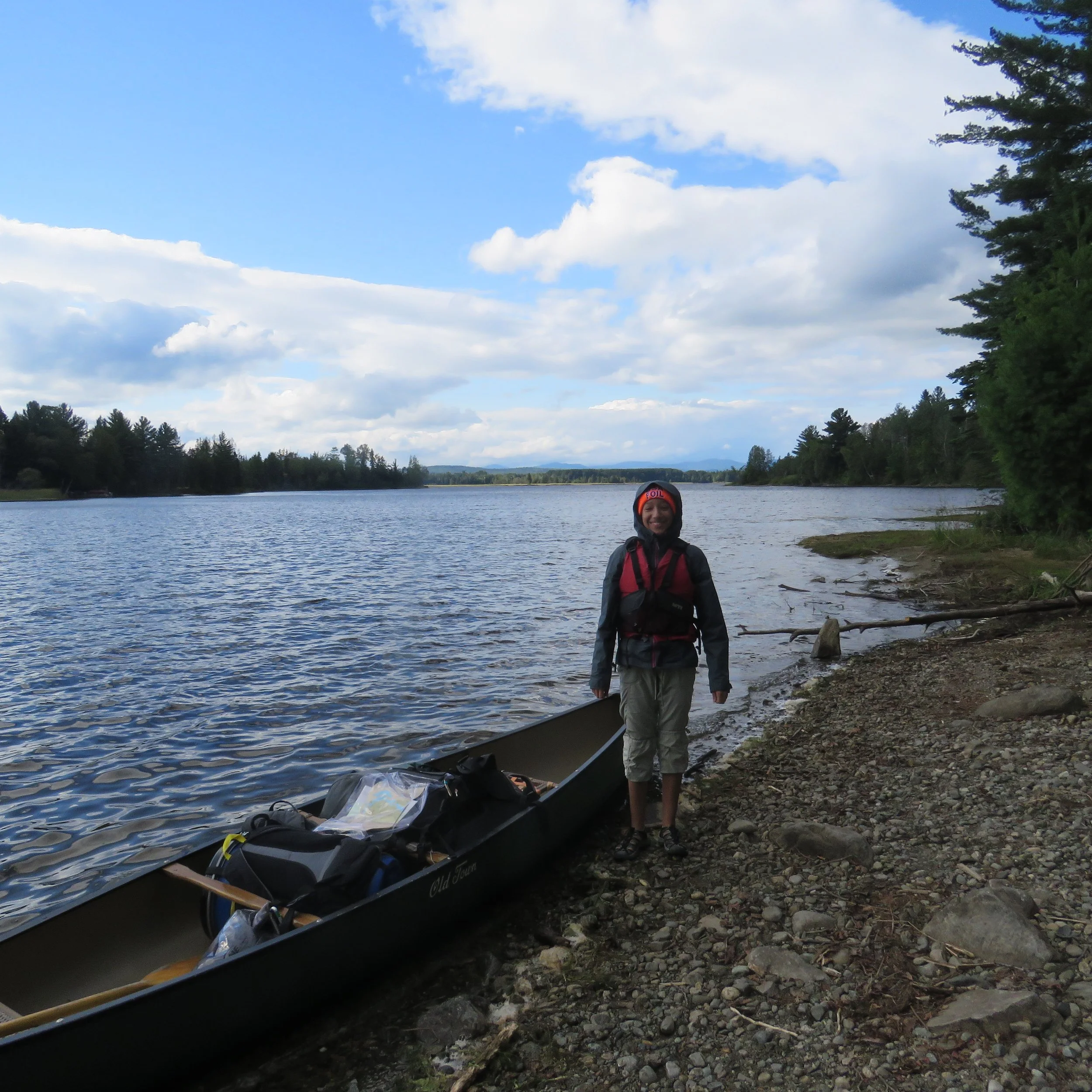 A person standing on a rocky shore next to a canoe on a river with a forested landscape and a partly cloudy sky in the background.