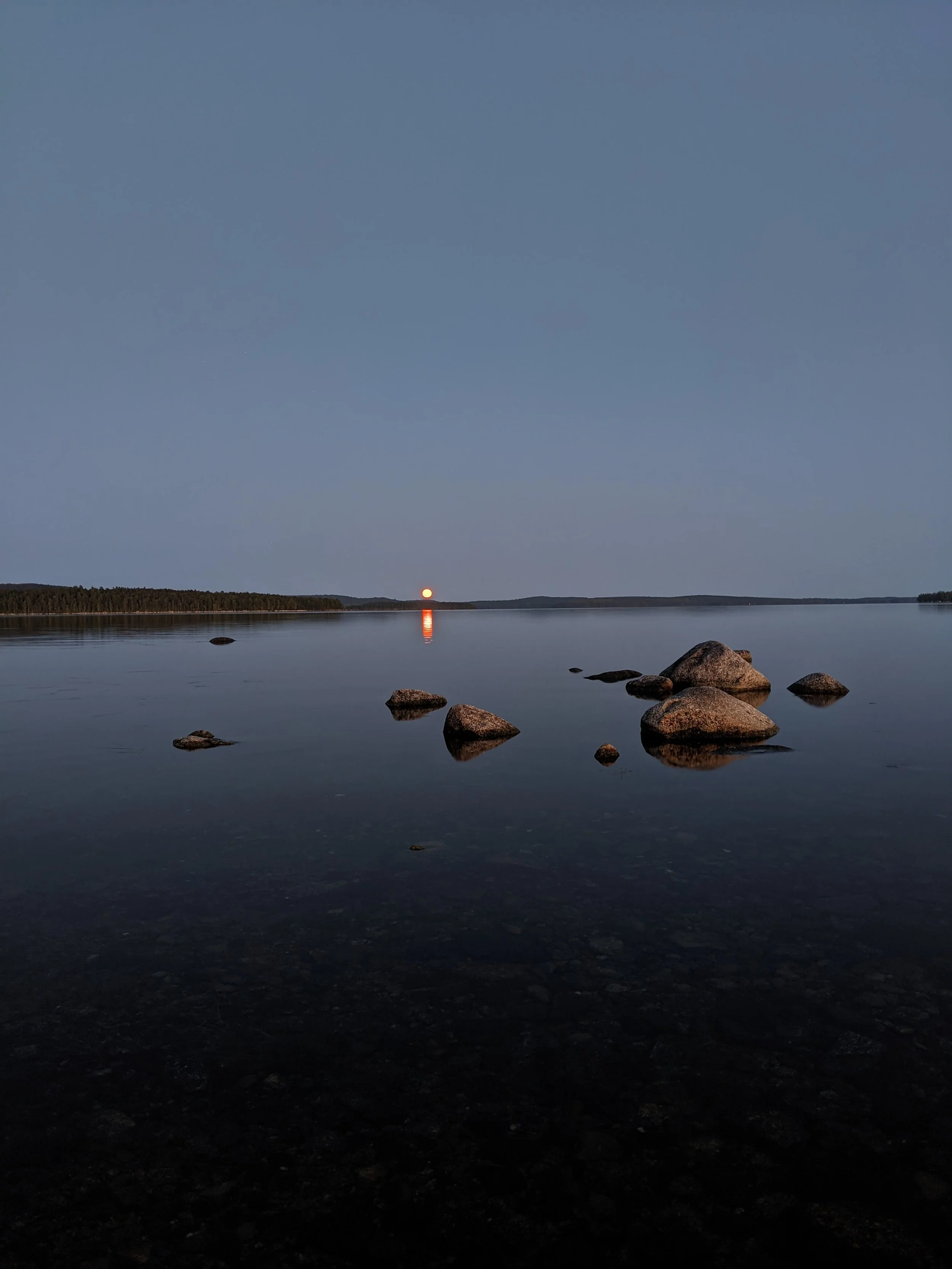 West Grand Lake, Maine. A calm body of water at dusk with rocks in the foreground, a forested shoreline on the horizon, and a bright orange moon rising or setting near the horizon reflection on the water.