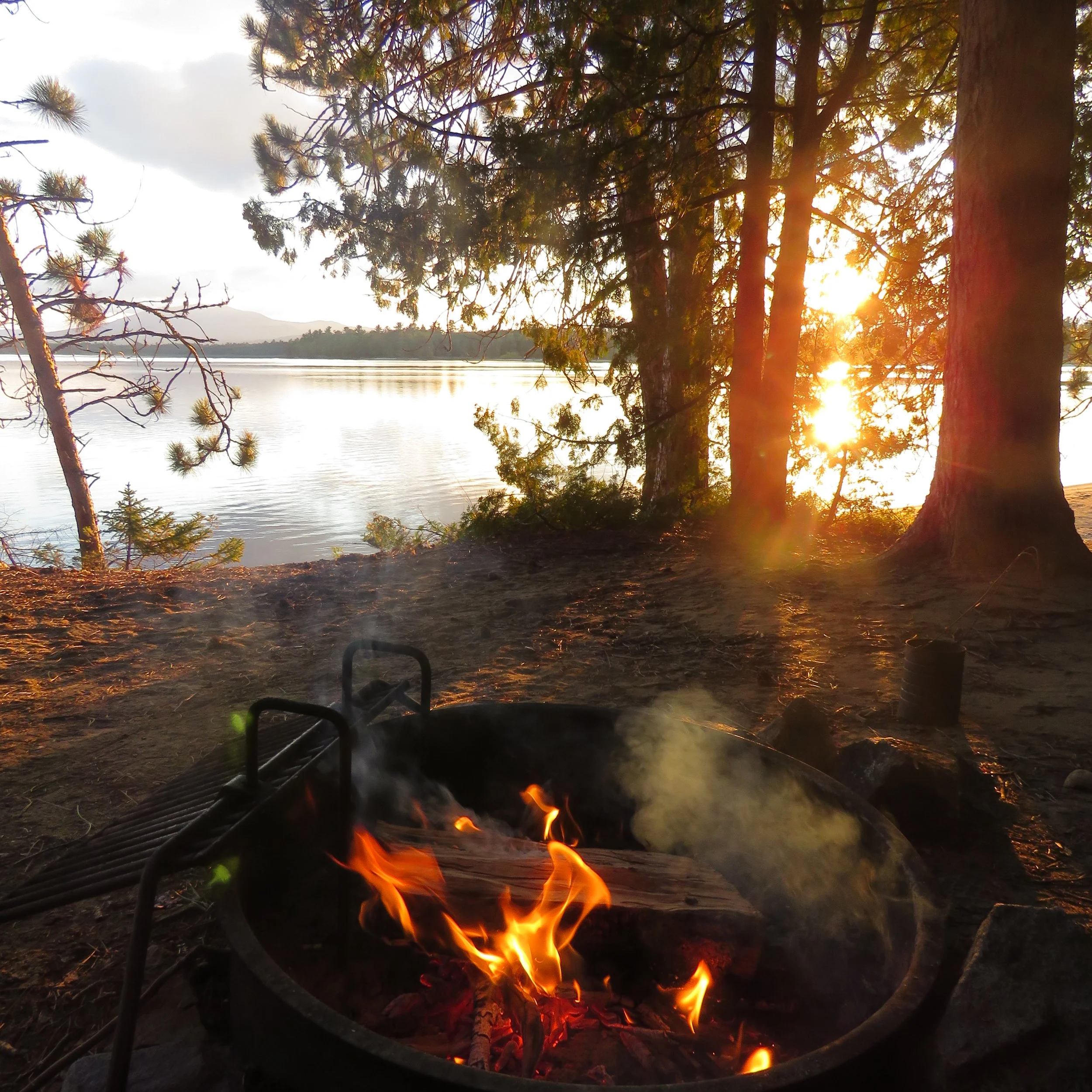 Campfire burning with visible flames and glowing embers in a forest by a lake at sunset, with trees and mountains in the background.