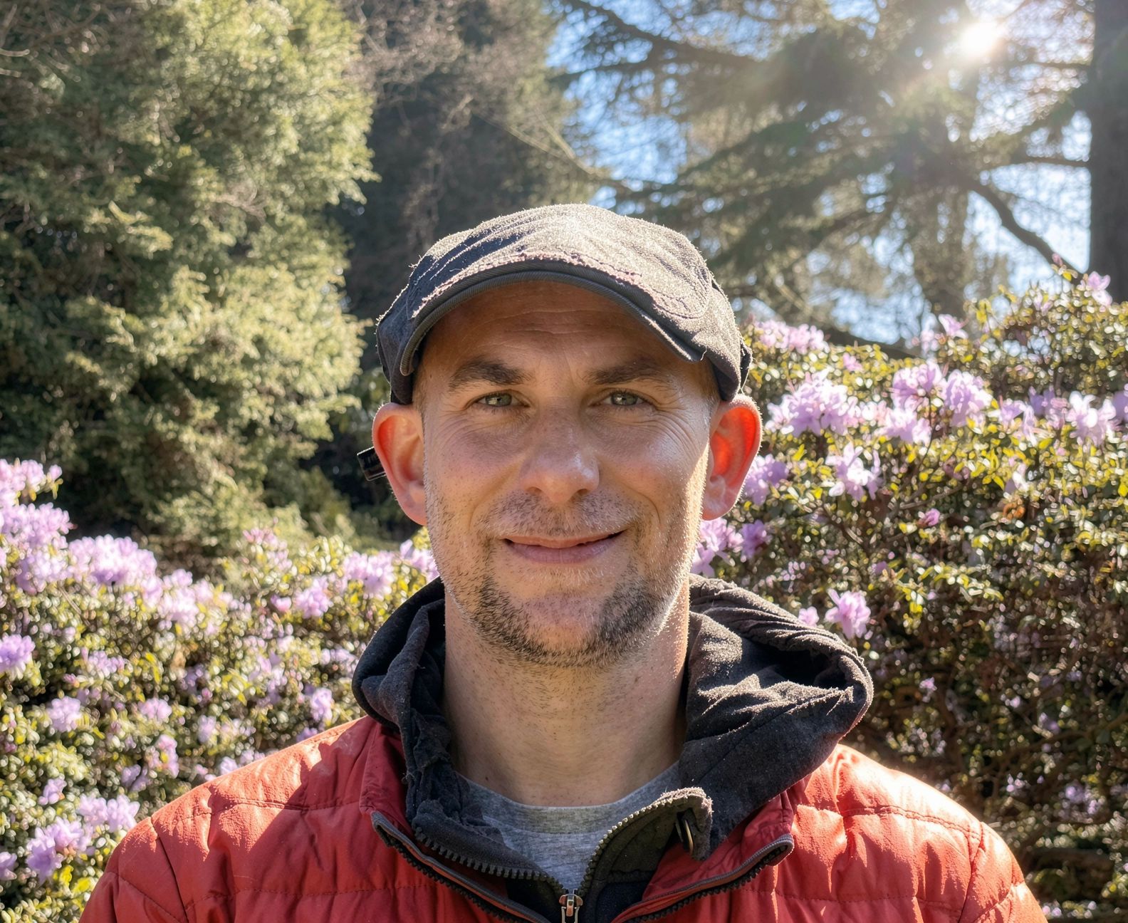 A man smiling outdoors in front of pink flowering bushes and trees, wearing a black cap and an orange jacket on a sunny day.