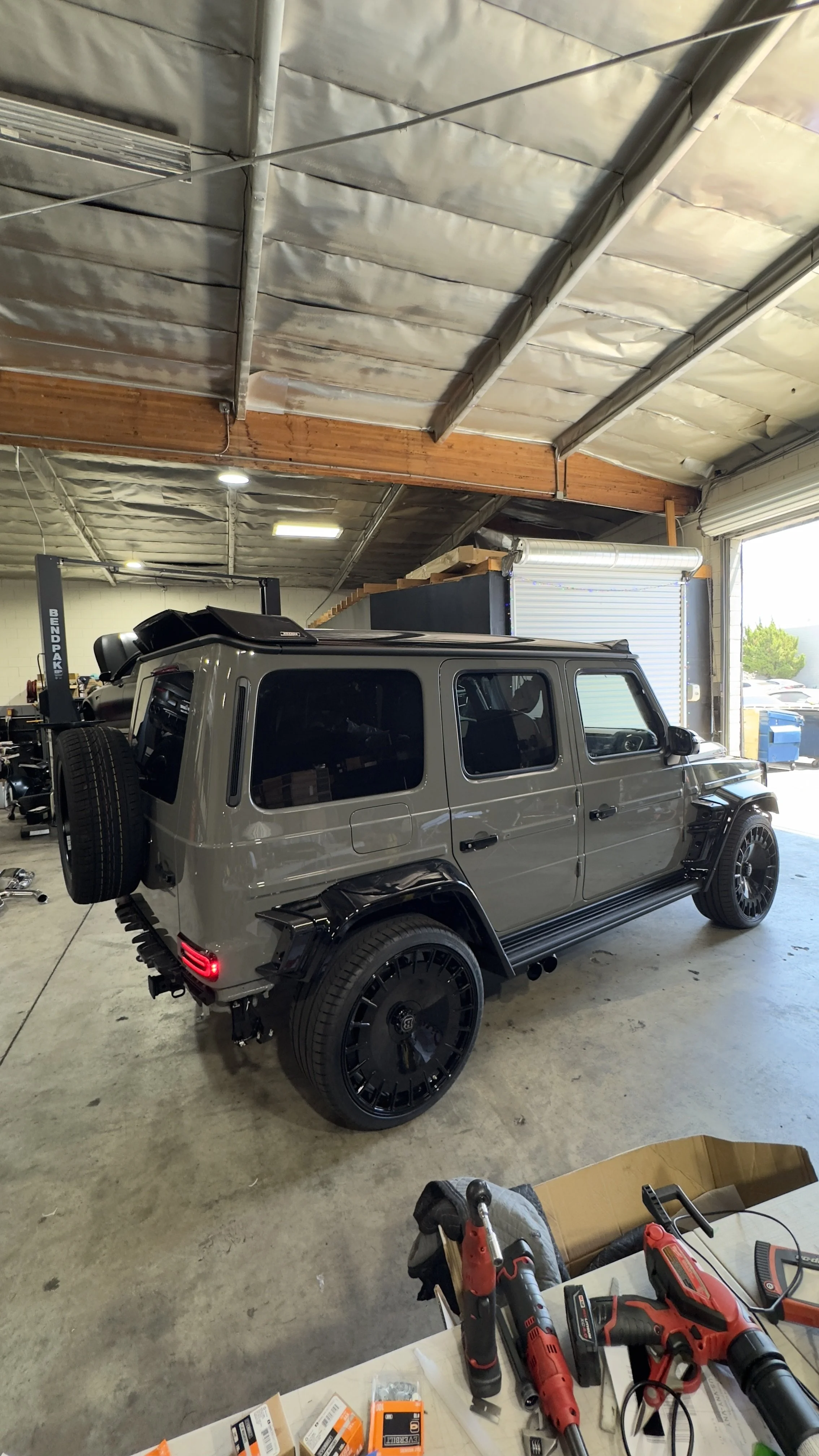 A gray 2026 Mercedes-Benz G63 with a body kit retro-fit, black upgraded wheels and a spare tire mounted on the back, parked inside a garage with an open roll-up door and some tools on a table in the foreground.