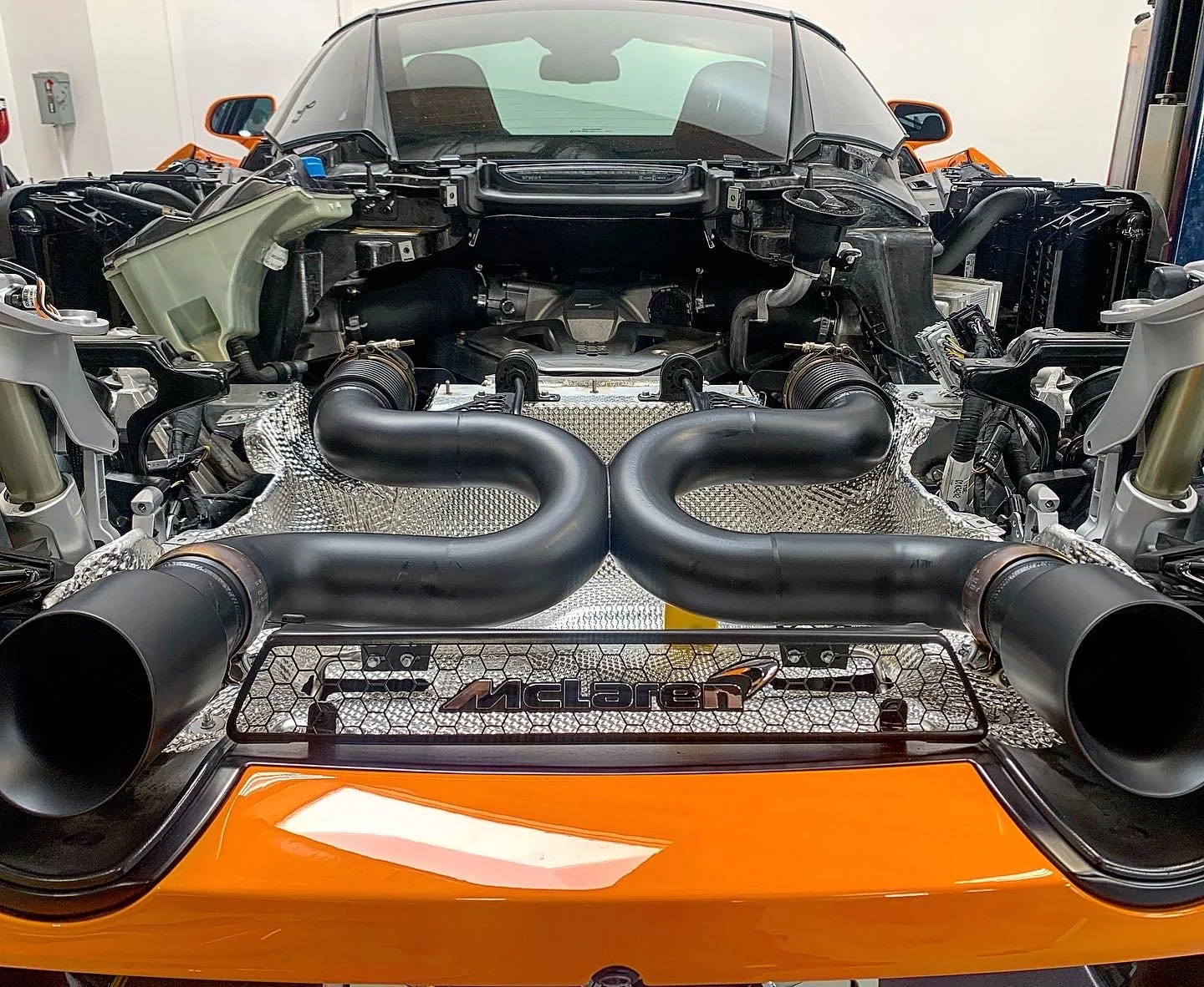 Close-up of a McLaren 720s with the rear dissembled, exposing the engine and exhaust system. The engine and gearbox bay, revealing a black exhaust pipes system from Voodoo and various mechanical parts, with the McLaren logo visible on a mesh grille.