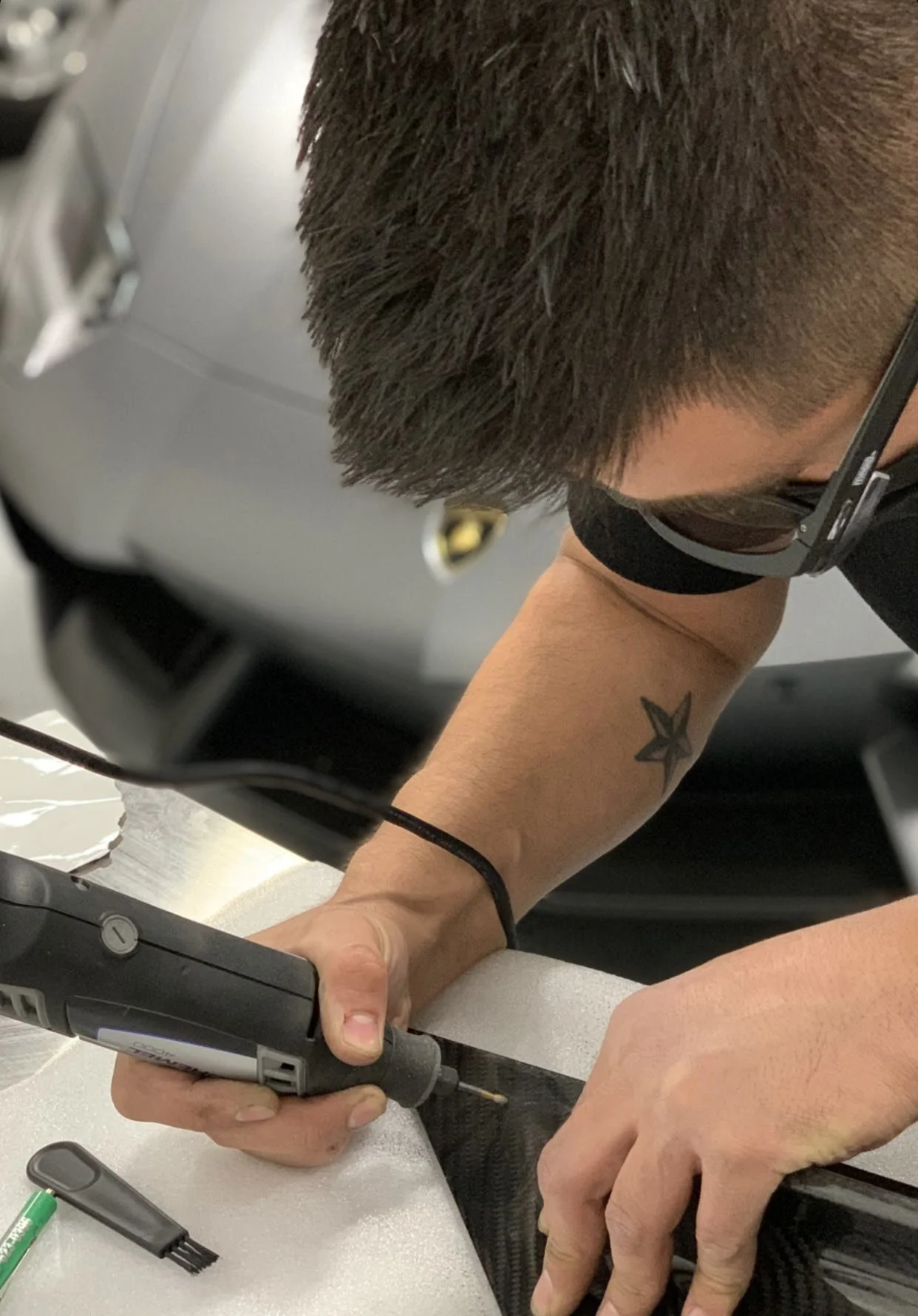 Cesario is trimming a carbon fiber panel, for a Lamborghini Aventador, to get the best body panel fitment possible, in a workshop. He has a star tattoo on his arm, wears eye protection glasses and a black mask, and is focused on the task.