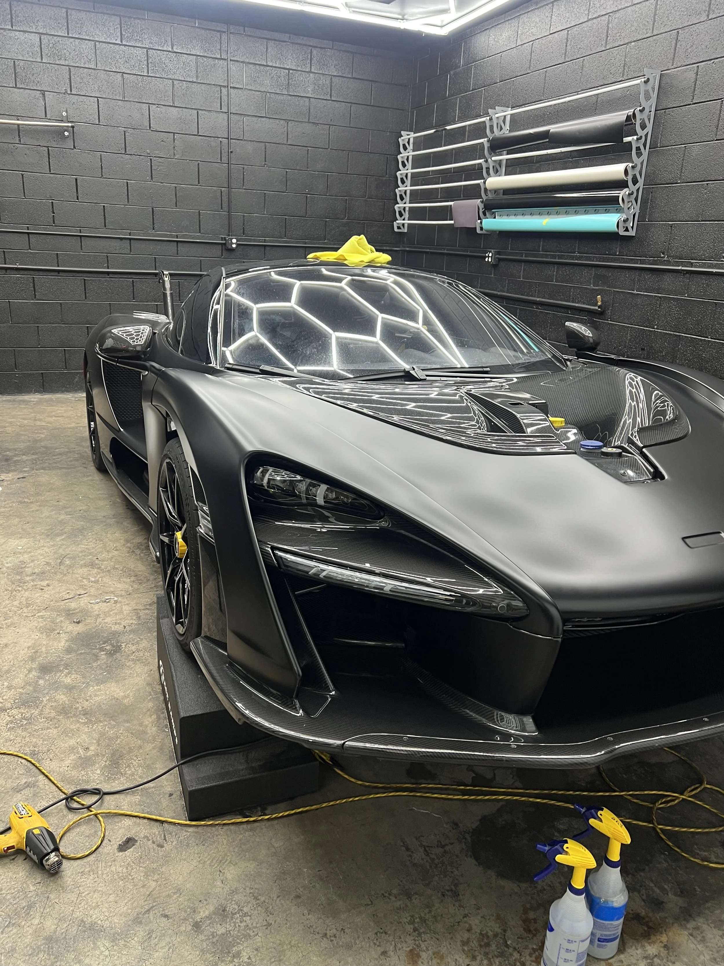 A McLaren Senna with Satin PPF installed. The car's hood and windshield are covered with protective film, reflecting hexagonal light fixtures overhead. On the wall behind, there are multiple rolls of wrapping or tinting film on a rack. Tools on floor