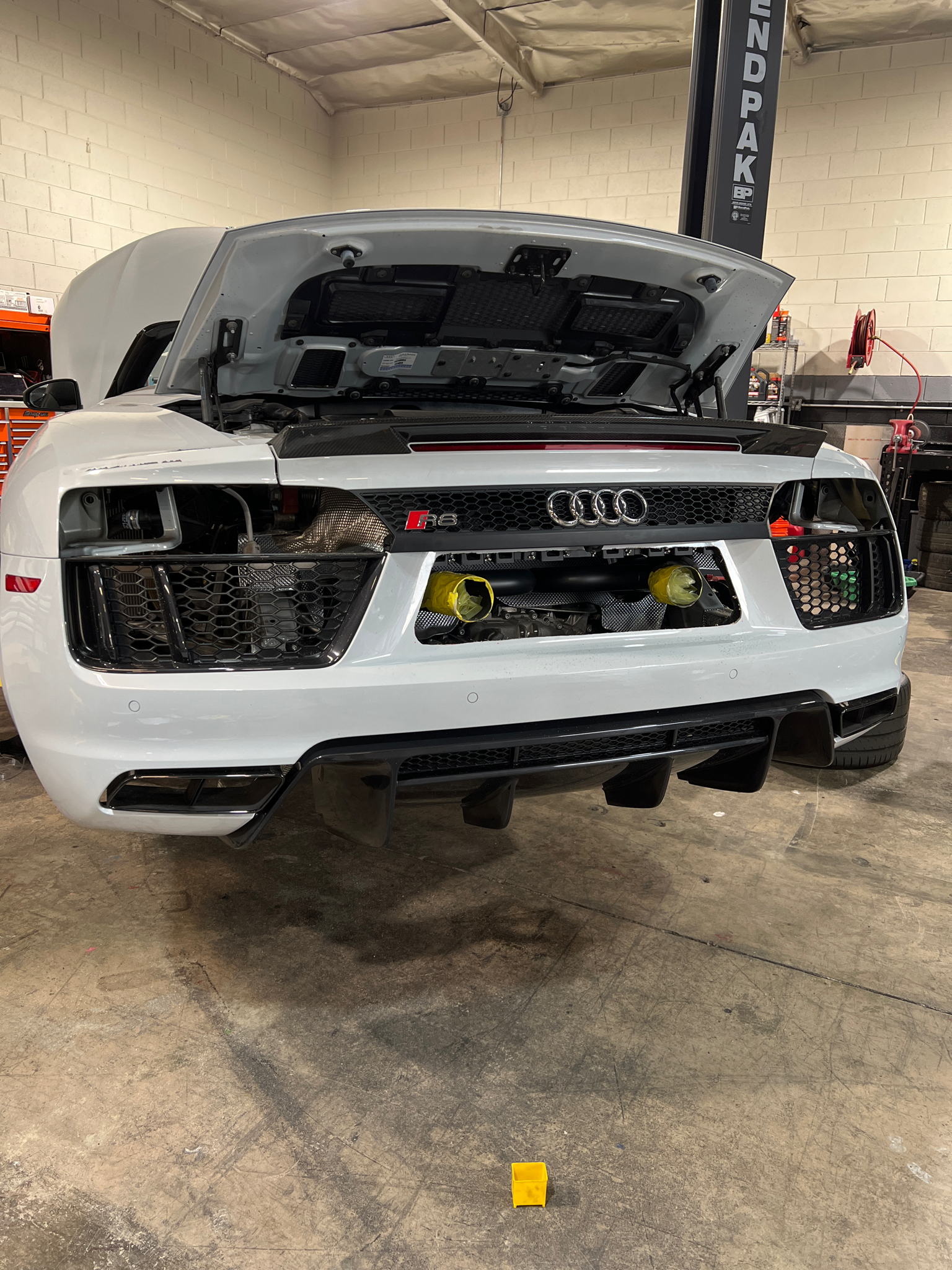 Rear view of a white Audi R8 Spyder in a workshop with the bonnet open, showing the engine compartment and rear exit exhaust retro-fit from Voodoo engineering.