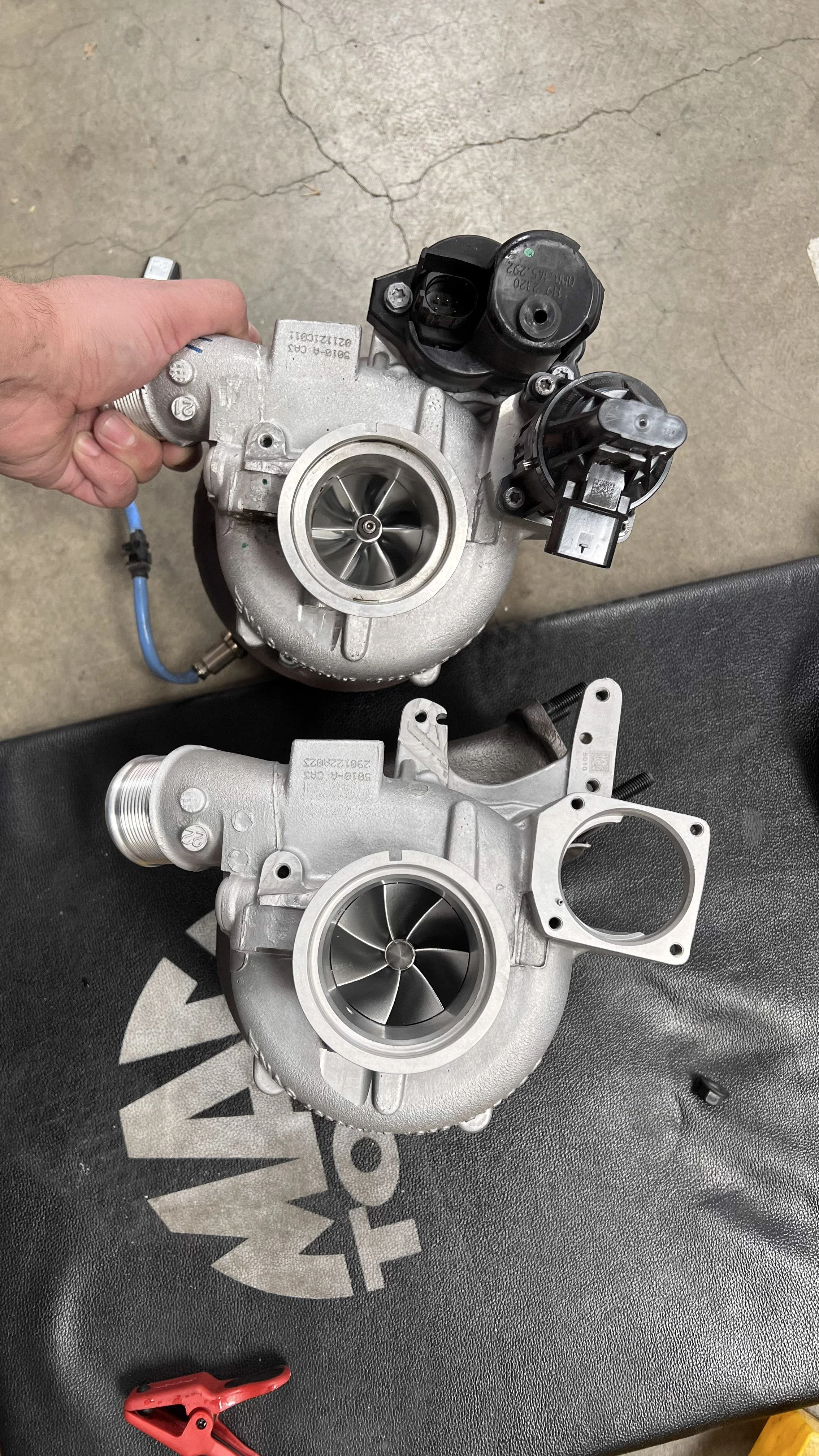 Two turbochargers for a Porsche Turbos S 992, the factory turbo being held by a Cesario's hand, placed on a black work surface with a red clamp and a concrete floor background. The other turbo not being held is an upgraded larger Pure Turbo. 