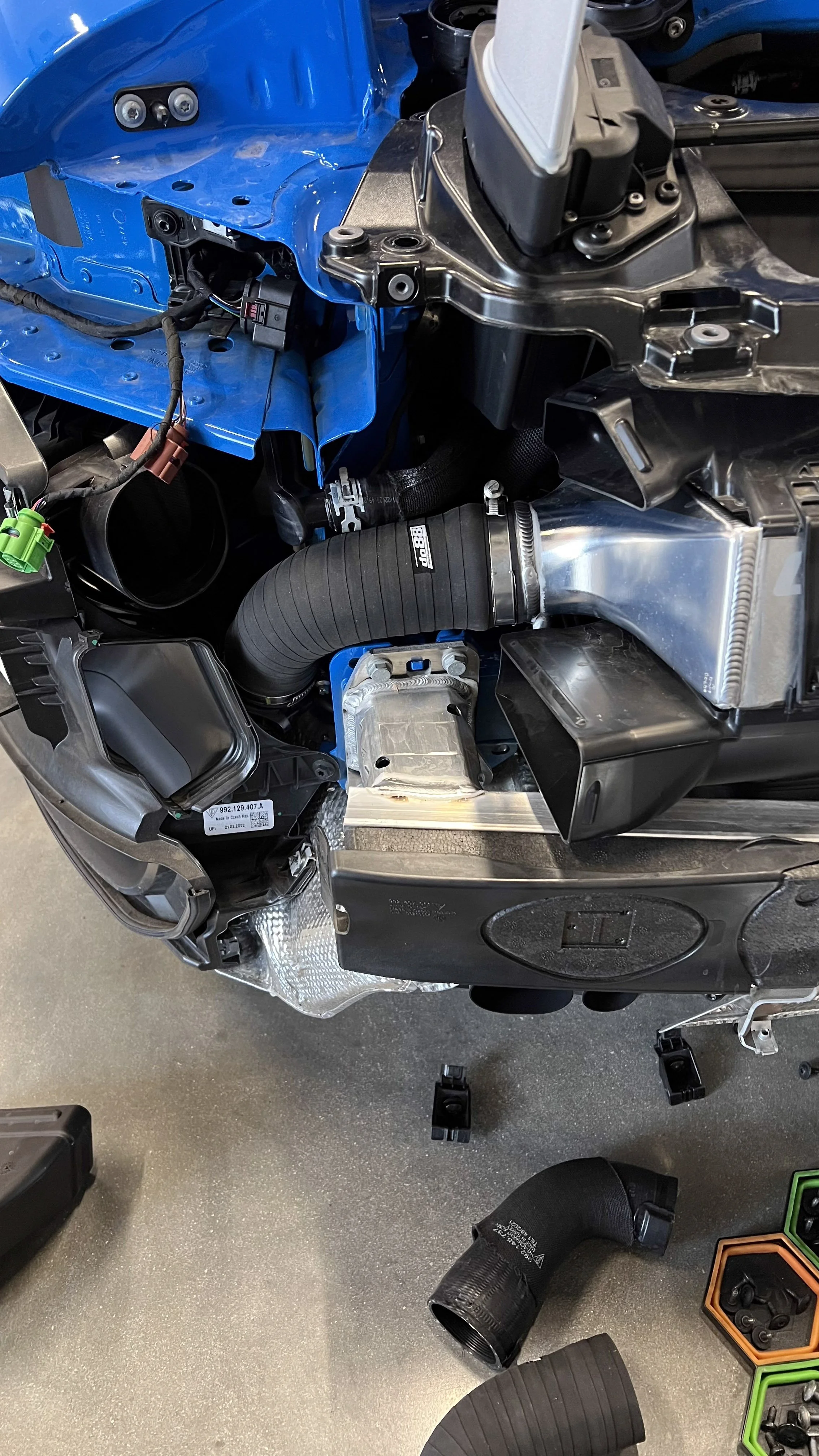 Close-up of a Porsche Turbo S 992 backend with newly retro-fitted CFM intercooler with various components, hoses, and wiring, with some parts unassembled and tools nearby.