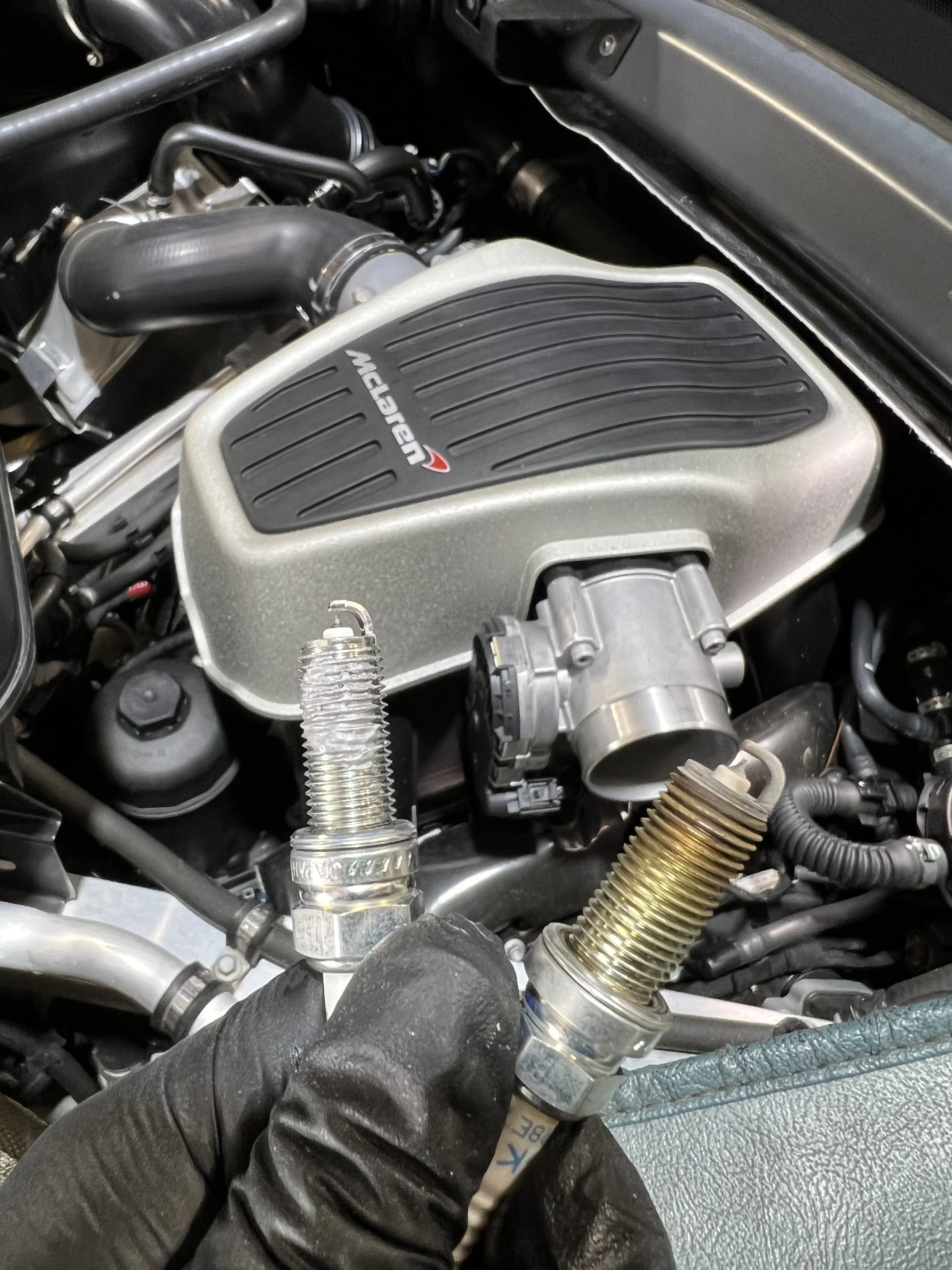 The master mechanic Cesario holding two spark plug. One worn and one new replacement in front of a McLaren 600LT engine.