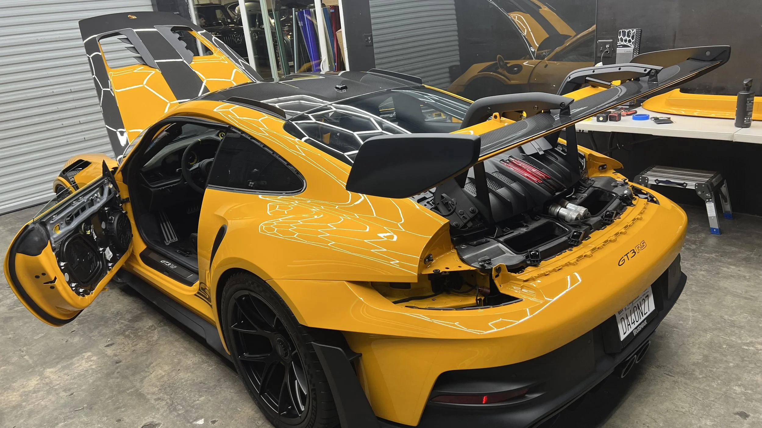 Yellow Porsche 911 GT3 RS getting ready for PPF install with front hood open and rear wing installed, inside a garage with tools and equipment on a workbench.