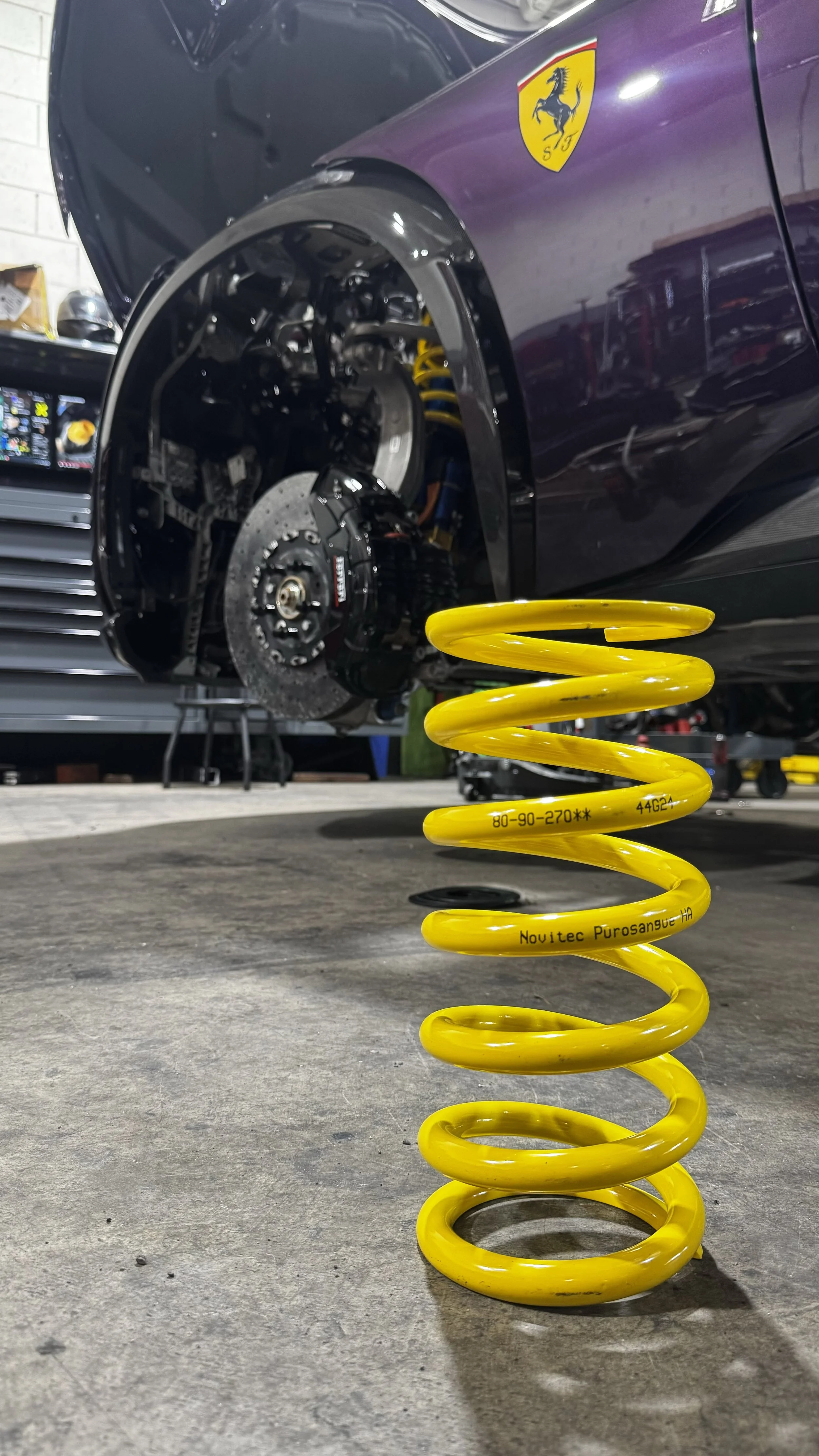 Yellow lowering coil spring from Novitec in front of a purple Ferrari Purosangue sports car with an open wheel well, exposing suspension and brake components in a garage or workshop.