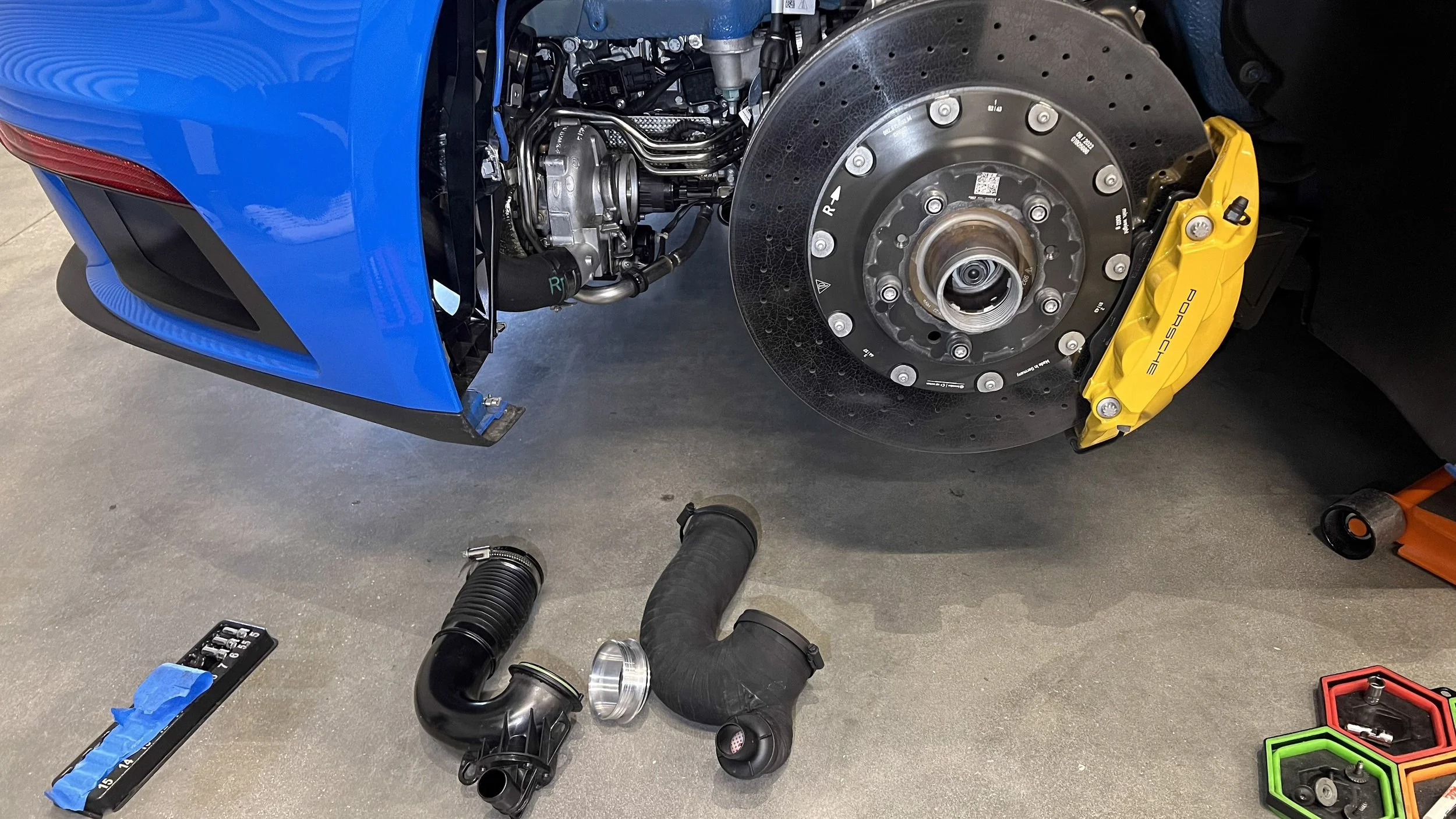 A blue Porsche Turbo S 992 sports car with the rear wheel removed, exposing the brake rotor and yellow brake caliper. Various car parts and tools are on the floor, including black turbo hoses, a tool with blue tape, and colorful hexagonal containers.