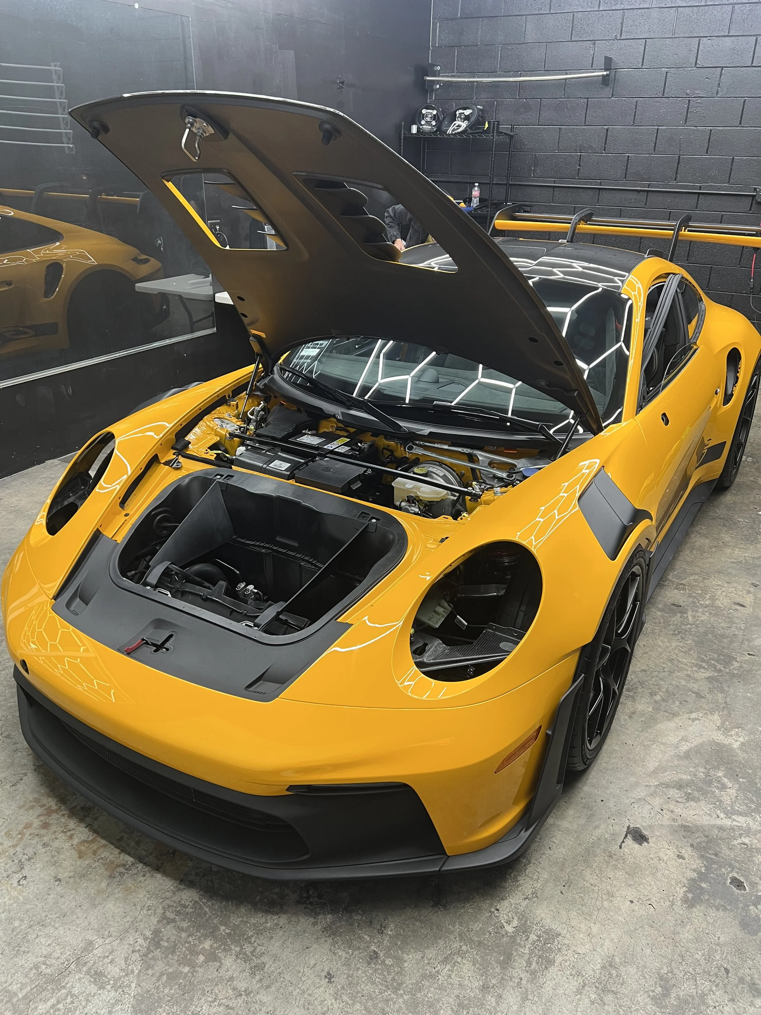 Yellow Porsche GT3rs sports car with hood raised and headlights removed in a PPF garage, showing trunk compartment.