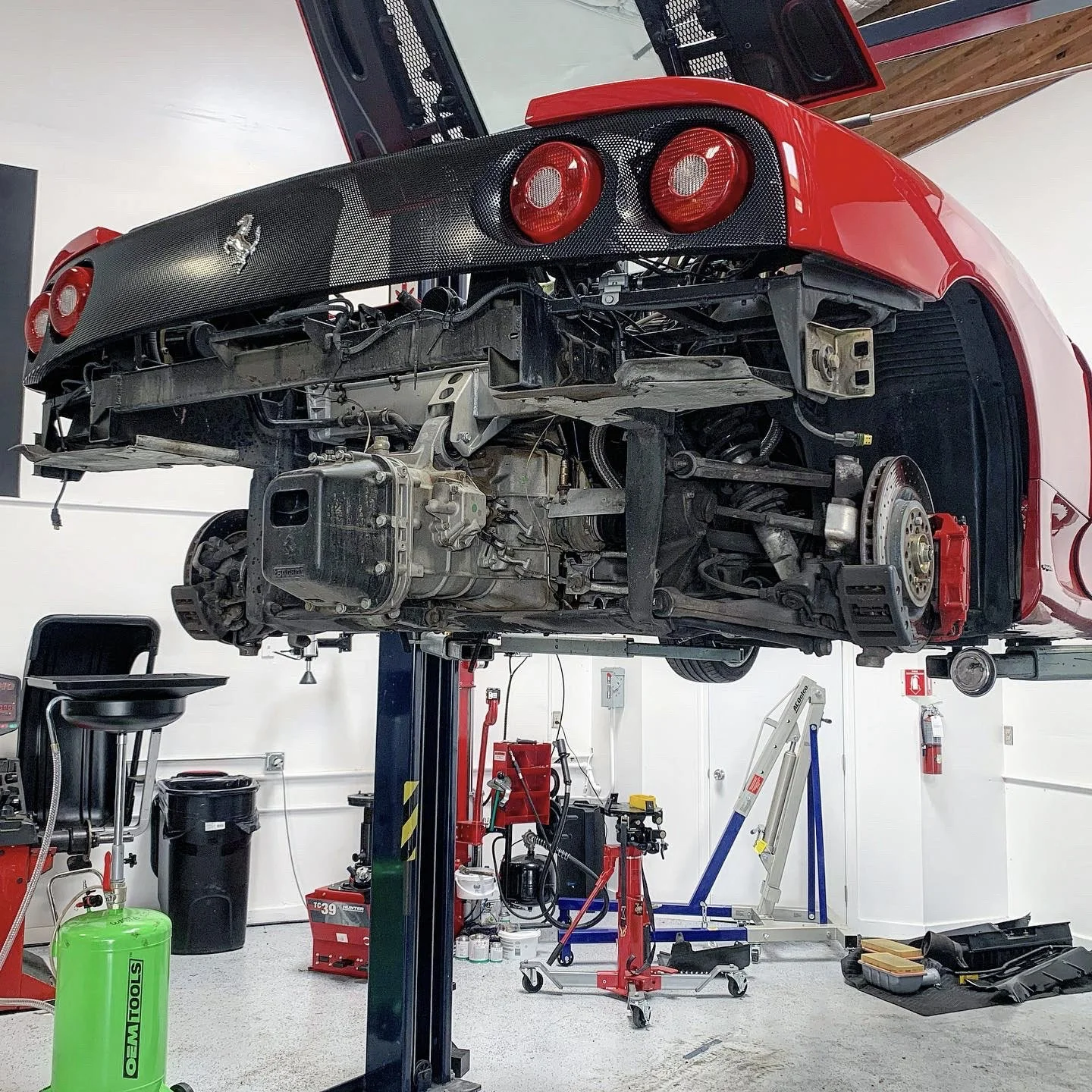 A Ferrari sports car is lifted on a hoist in an auto repair shop with its underside exposed, showing the engine and mechanical components.