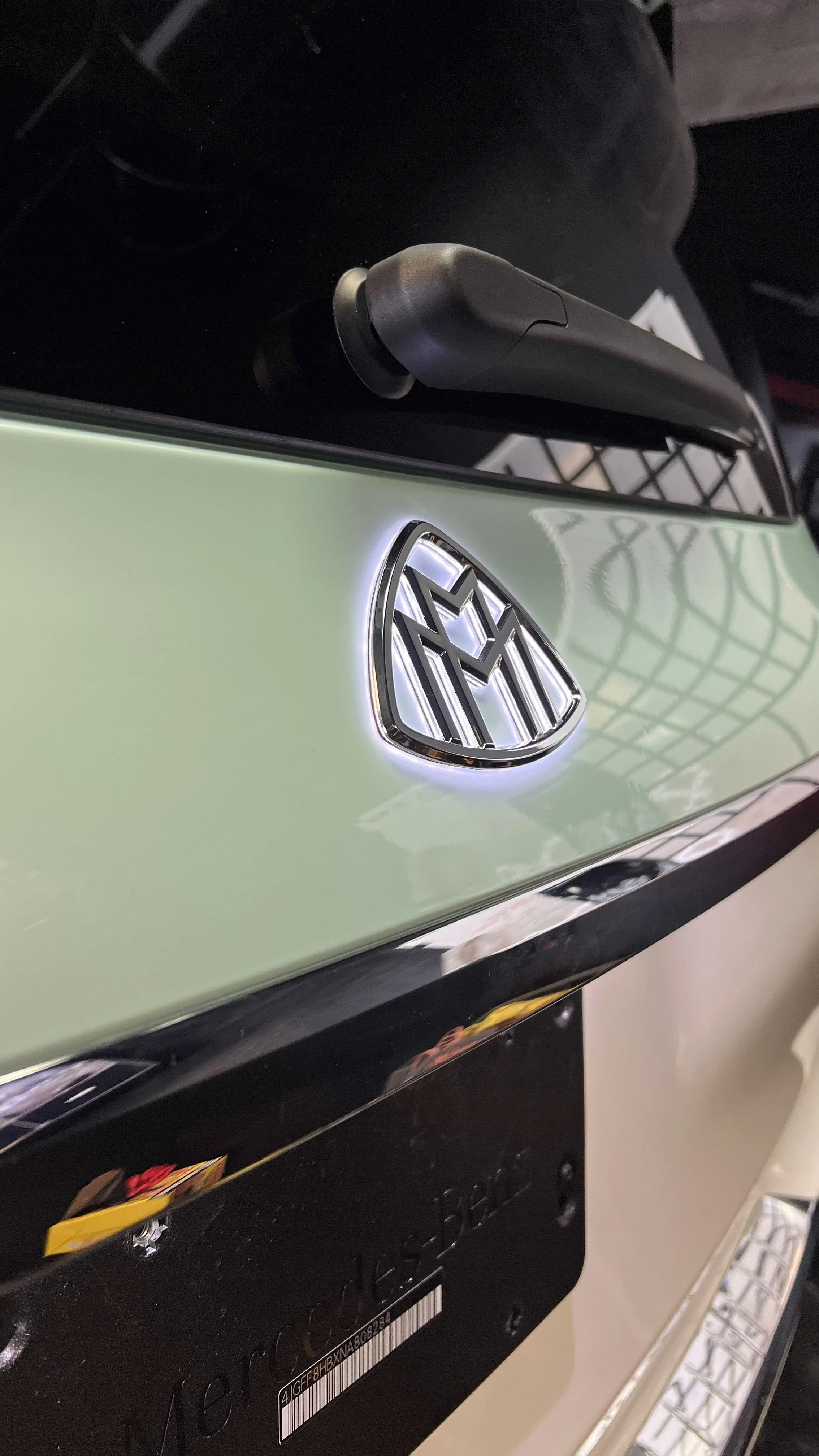 Close-up of a Mercedes-Benz GLS 580 Maybach luxury car trunk showing a retro-fitted Mercedes-Benz logo illuminated above a license plate holder.