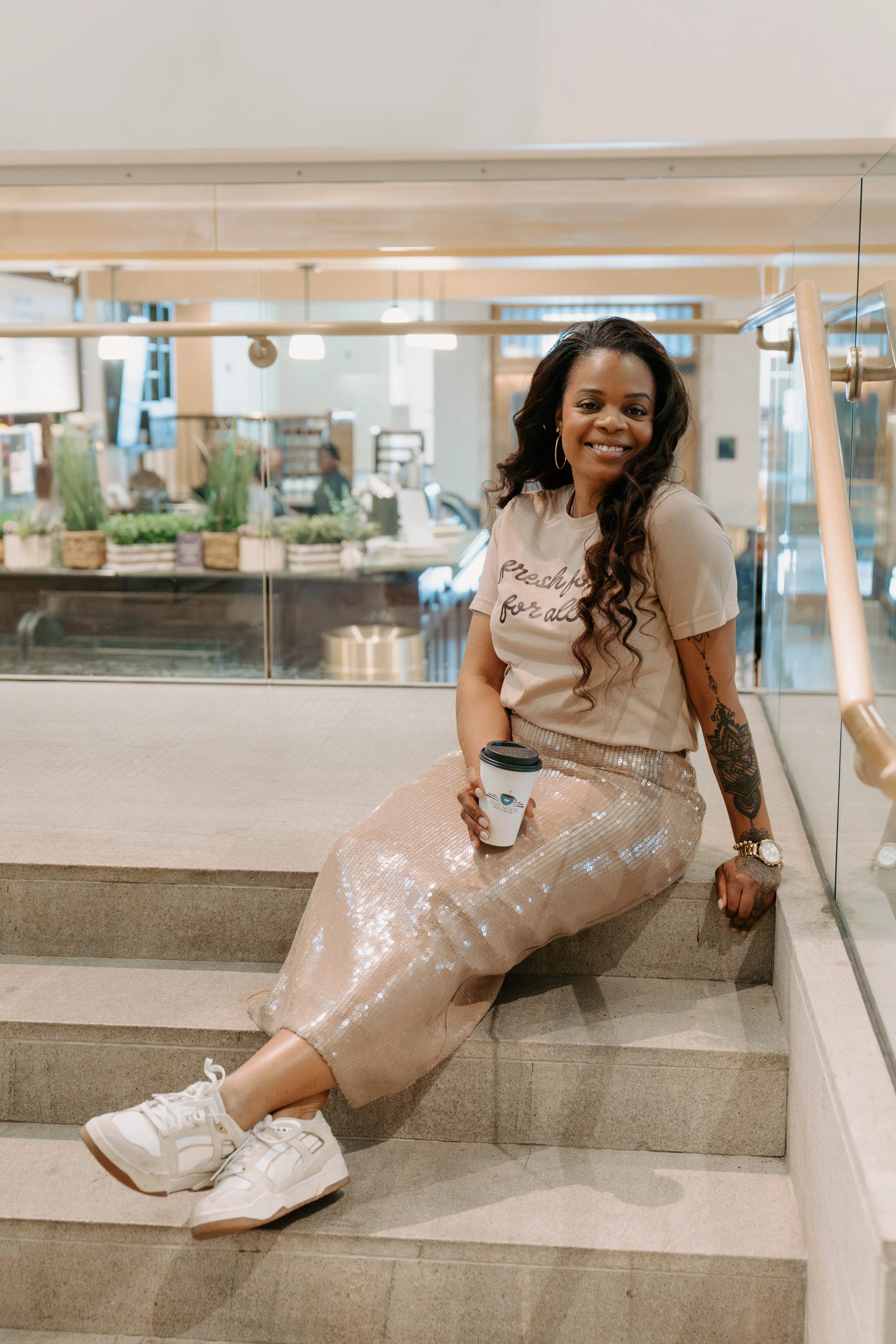 A woman with long dark curly hair, hoop earrings, and a watch sitting on stairs in an indoor modern space, holding a white paper coffee cup, smiling, wearing a t-shirt and a shiny beige skirt with sneakers.