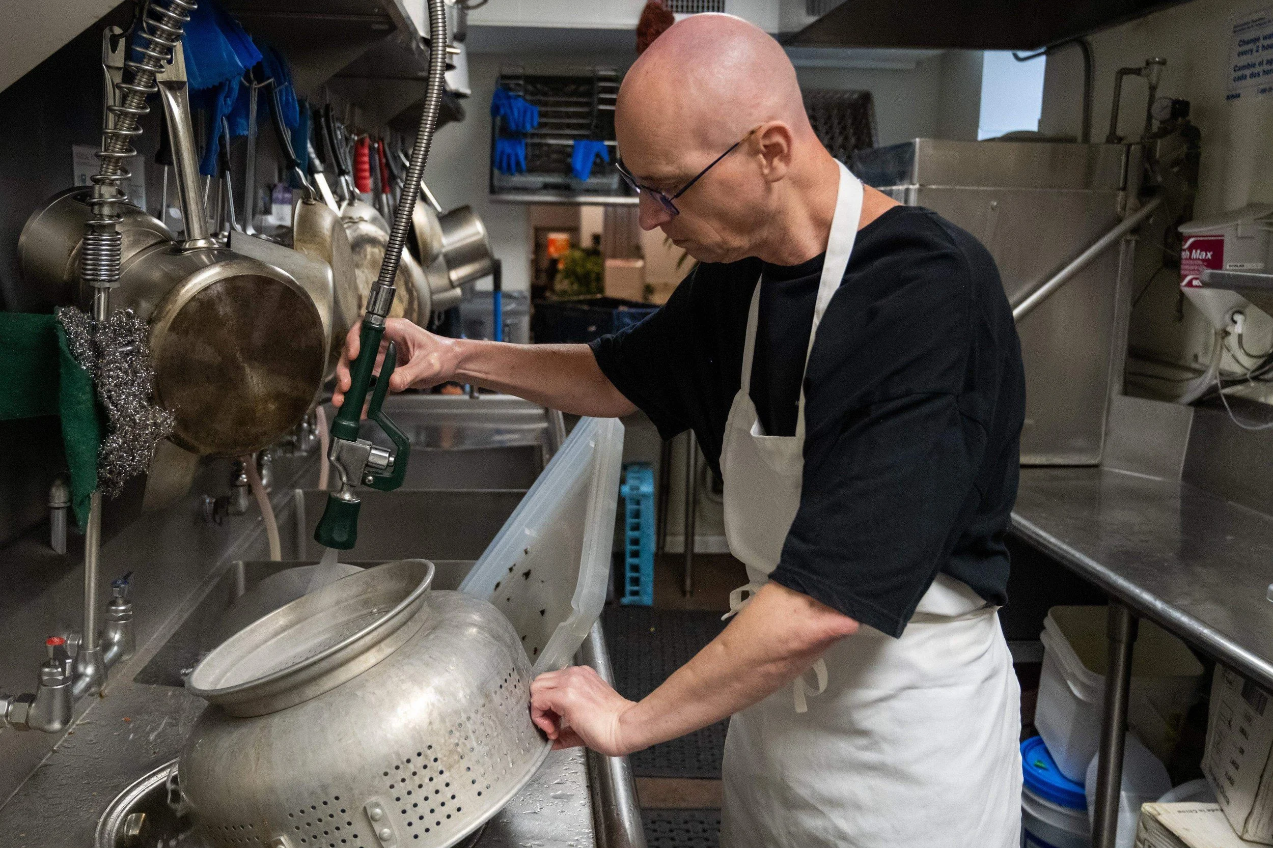A bald man wearing glasses, black shirt, and white apron working in a commercial kitchen with stainless steel utensils and equipment, focusing on a task involving a large metal container.