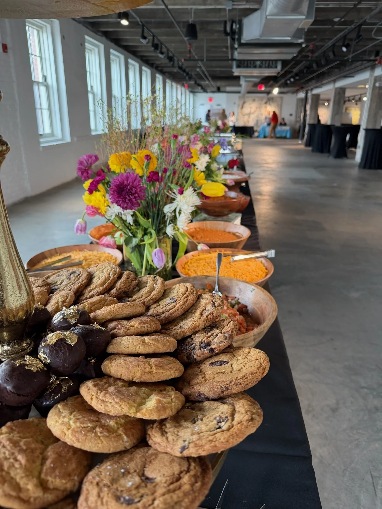 Table with assorted cookies, bowls of snacks, a floral centerpiece, and a buffet setup in a modern, industrial-style event space.