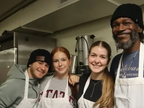 Four people smiling in a kitchen, wearing aprons and hats, standing close together.