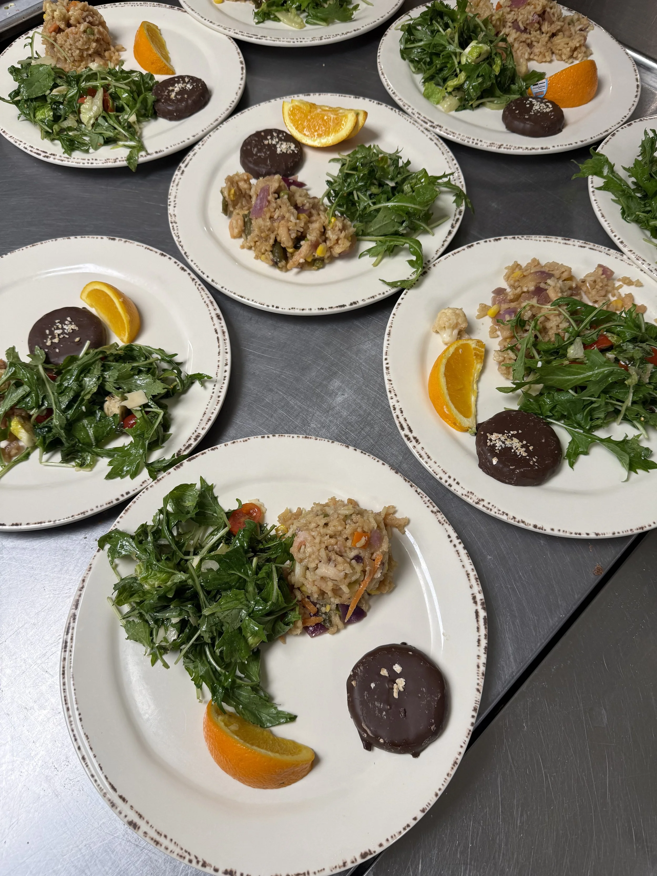 Multiple white plates with a salad, a scoop of rice mixed with vegetables and meat, a slice of orange, and a chocolate dessert with a sprinkle of salt, arranged on a stainless steel surface.