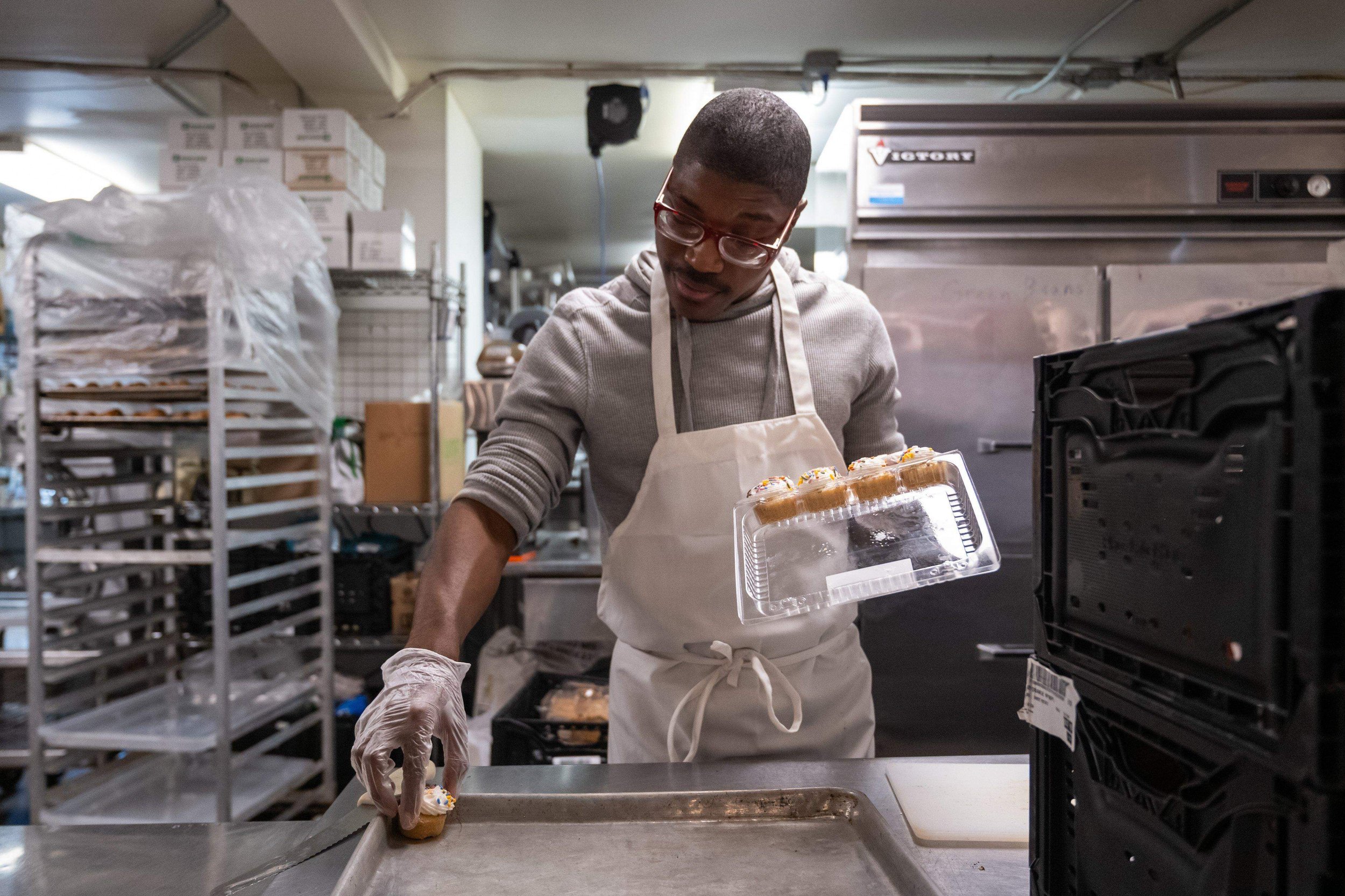 A man wearing glasses, a gray hoodie, and a white apron is working in a commercial kitchen. He is placing decorated cupcakes into a clear plastic container. The background shows metal shelves, boxes, and a large industrial refrigerator.