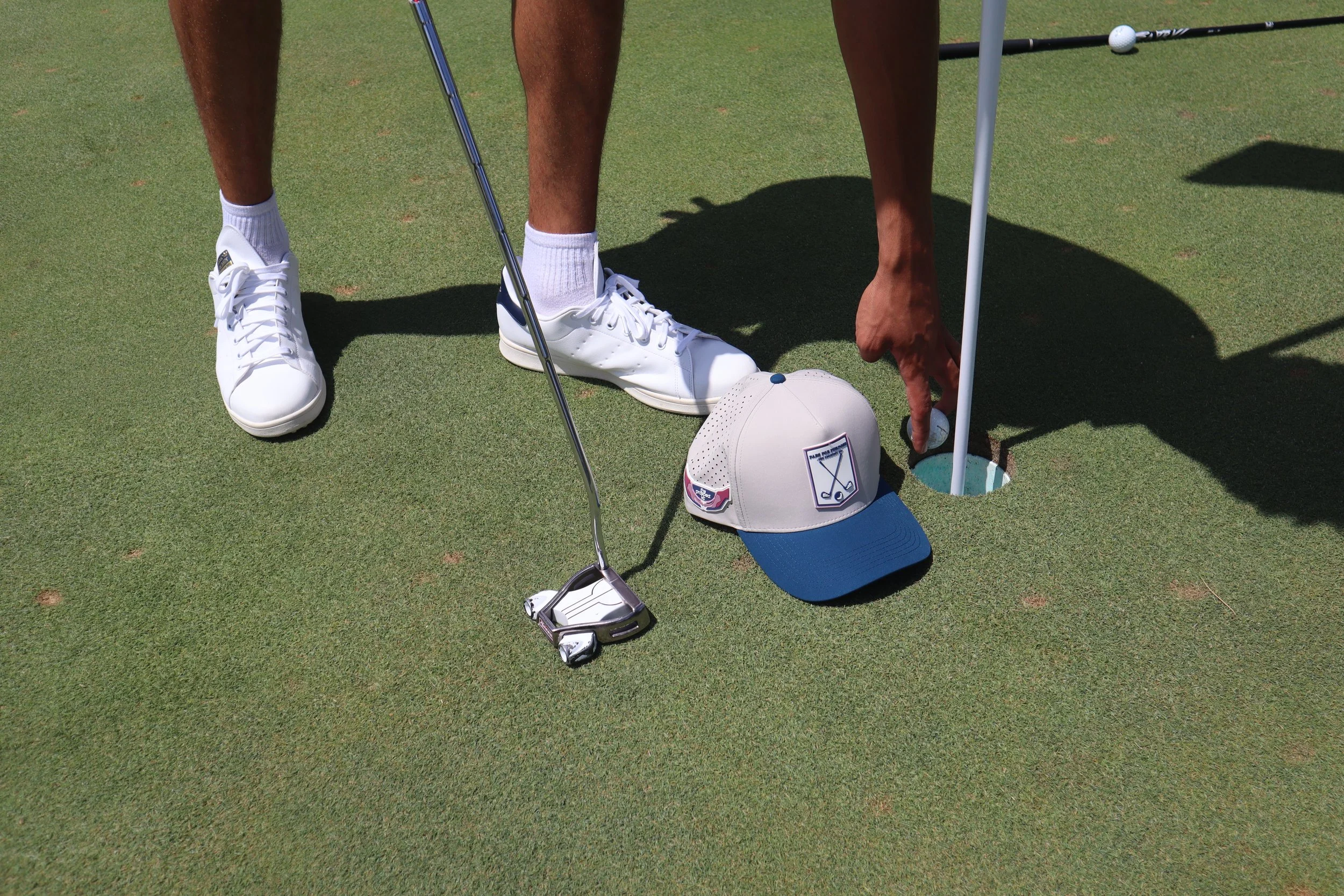 Close-up of a person preparing to putt on a golf green, with a golf club, golf ball, cap, and shoes visible.
