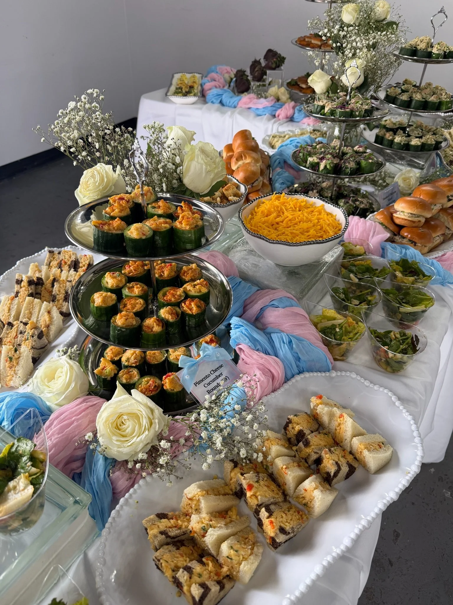 A table decorated with flowers and colorful cloth, displaying a variety of desserts including sandwiches, stuffed rolls, and other finger foods, arranged in trays and bowls for a celebration.