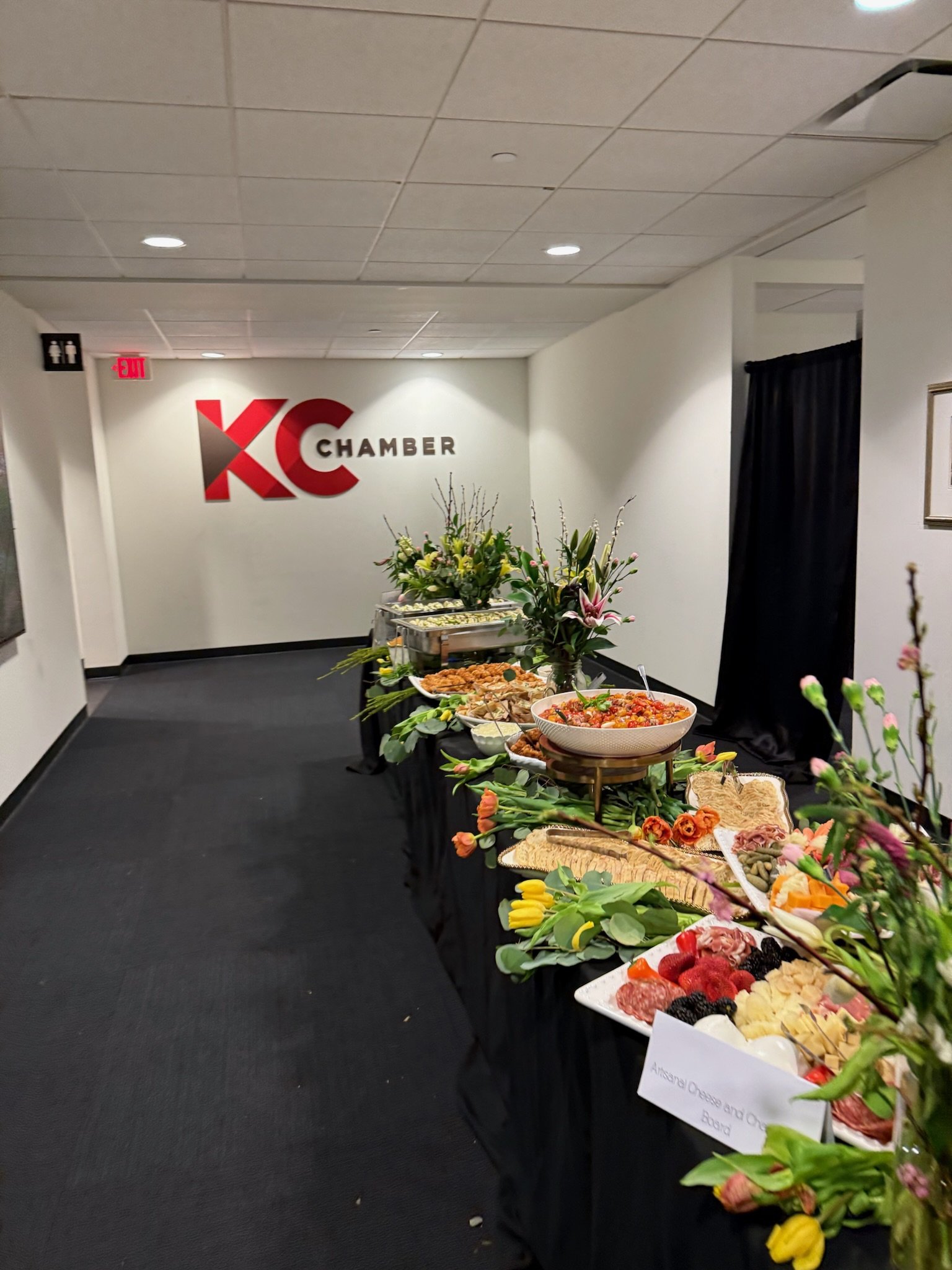 Buffet table with various appetizers and finger foods, floral arrangements in vases, in a room with a sign reading 'KC Chamber' on the wall.