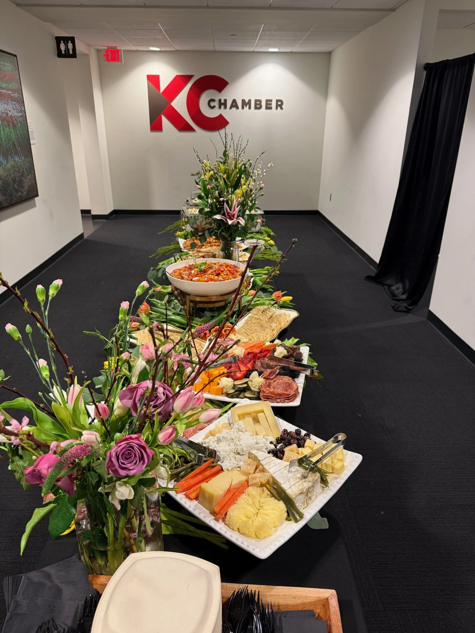 A buffet table with an assortment of cheeses, vegetables, and accompaniments, decorated with a large bouquet of pink and purple flowers, in an event space with a sign reading 'KC Chamber' on the wall.