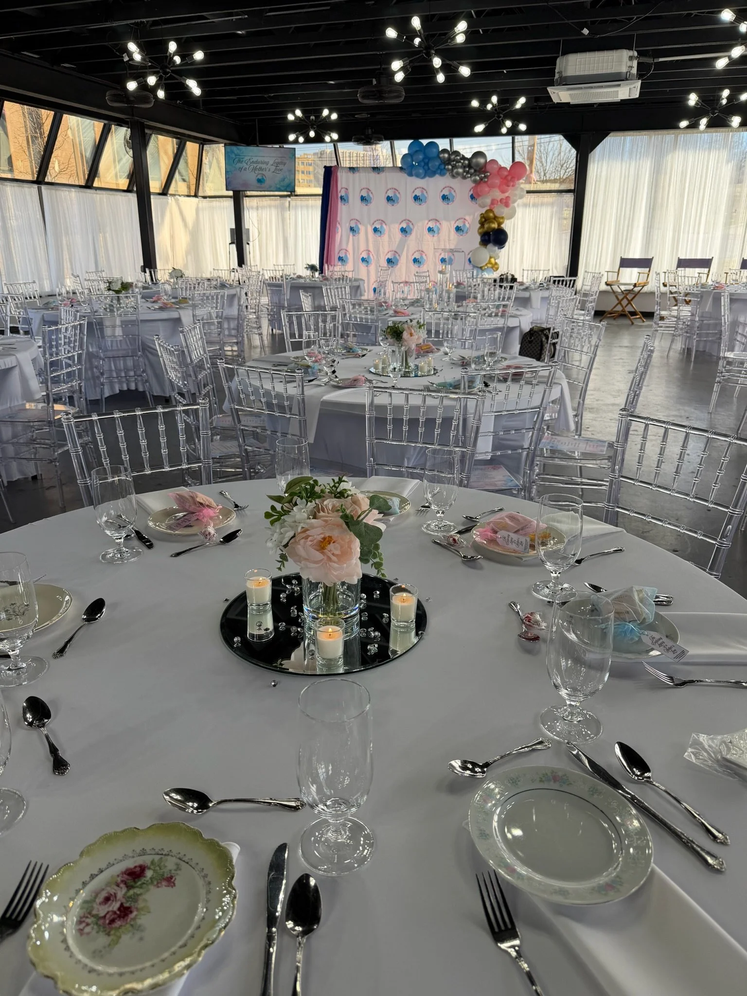 Elegant banquet hall decorated for a celebration with round tables, floral centerpieces, candles, and balloon arrangements on a pink, blue, gold, black, and white backdrop.