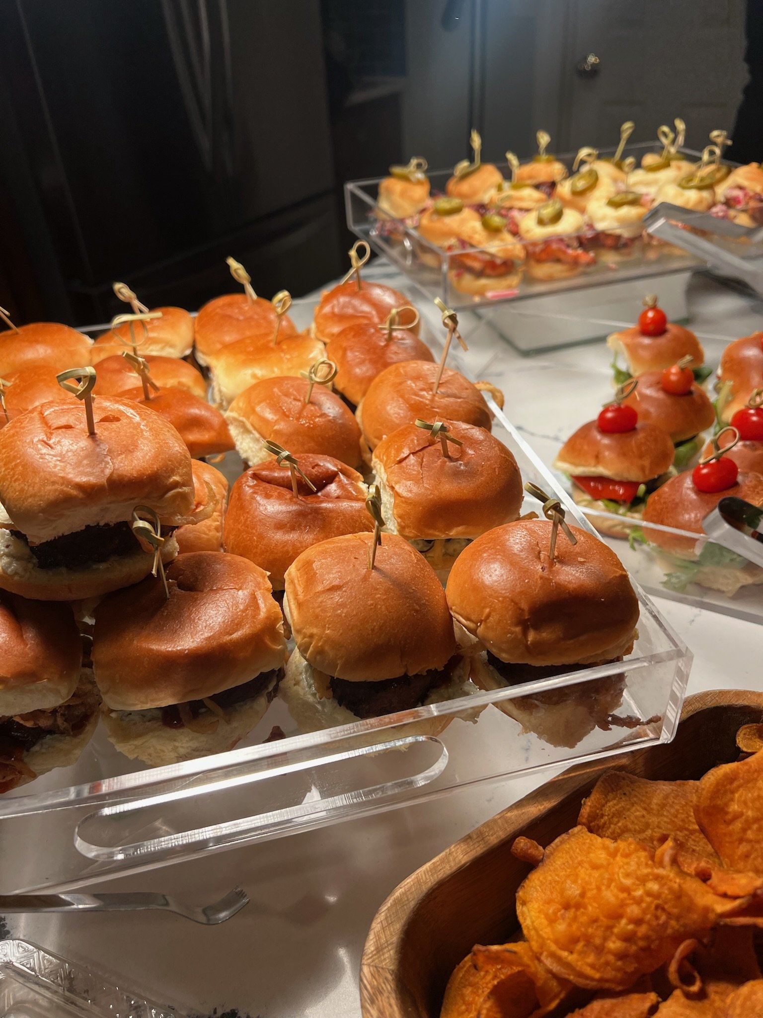 Assorted small sandwiches and sliders with toppings like pickles and cherry tomatoes, arranged on clear acrylic trays and wooden bowls.