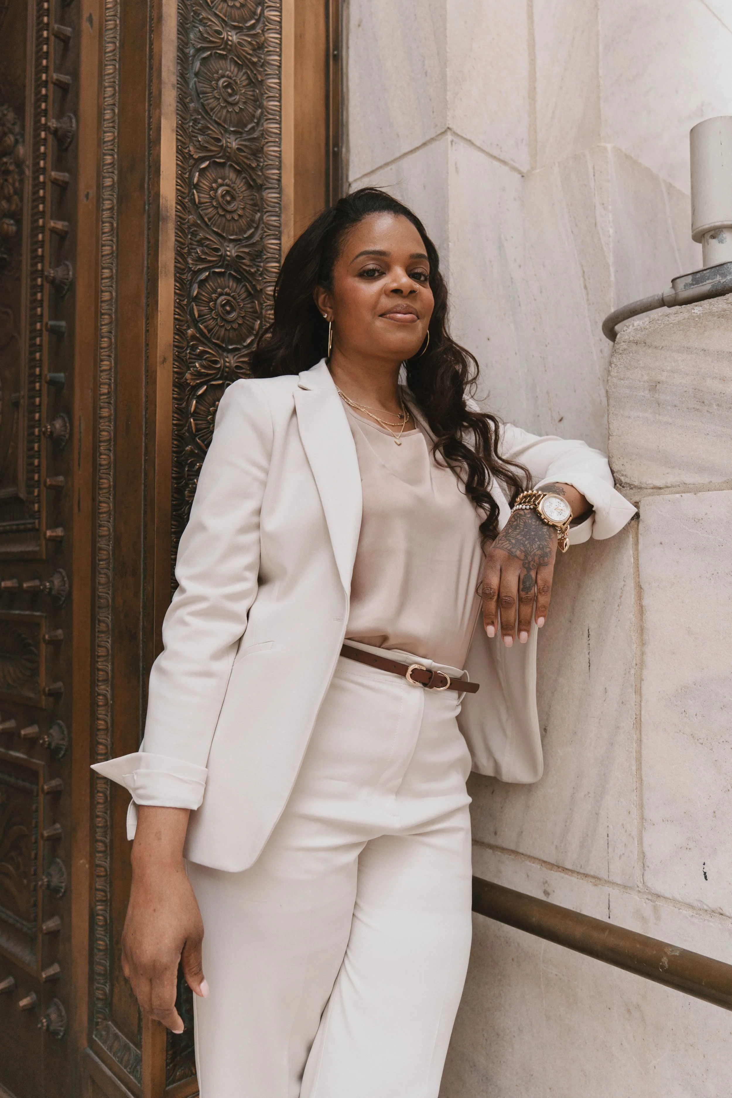 A woman in a white suit posing against a stone wall and decorative wooden door.