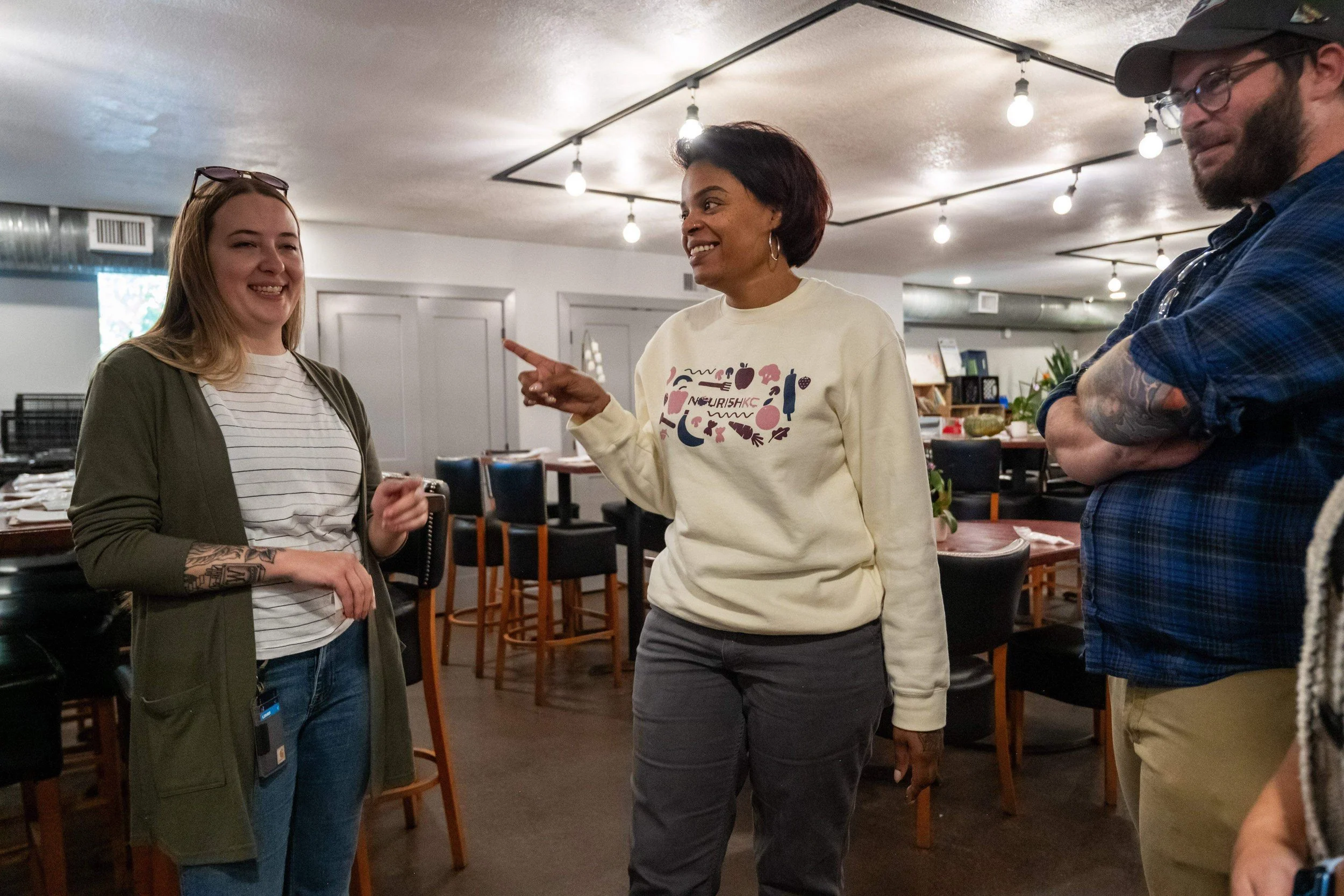 Three women are engaging in a conversation inside a restaurant or cafe, smiling and enjoying each other's company. The woman in the middle is pointing while talking, the woman on the left is holding a phone, and the woman on the right has tattoos on her arm and is wearing a cap and glasses.