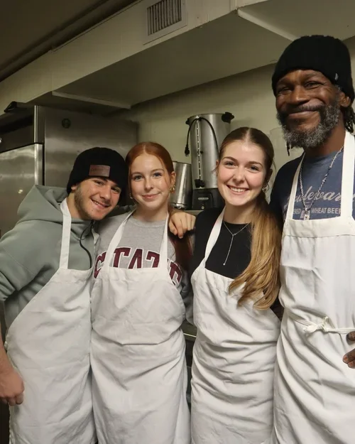 Four people wearing aprons smiling for a photo in a kitchen. Three are young women and one is an older man, all posing closely together.